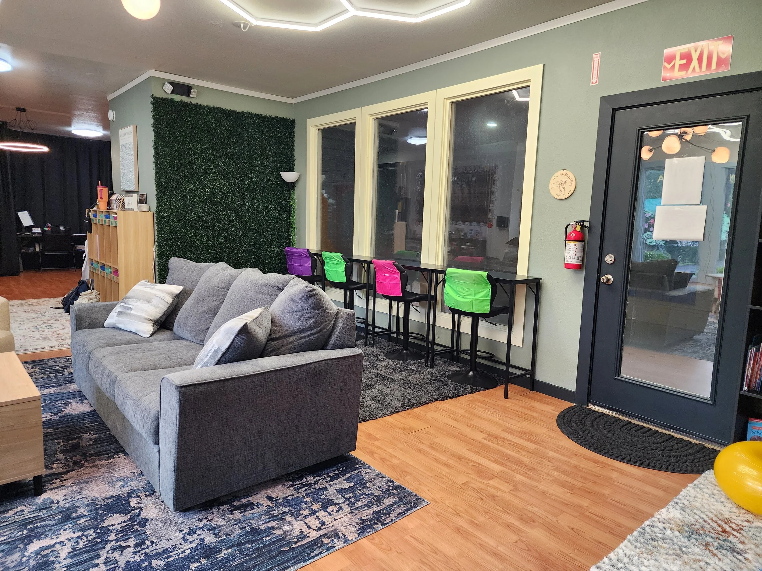Interior of a lounge or waiting area with a gray couch, colorful chairs, a green wall, and large windows. There is a black door with an EXIT sign and a fire extinguisher nearby. The wall behind the window has a decorative hedge panel and a standing lamp.