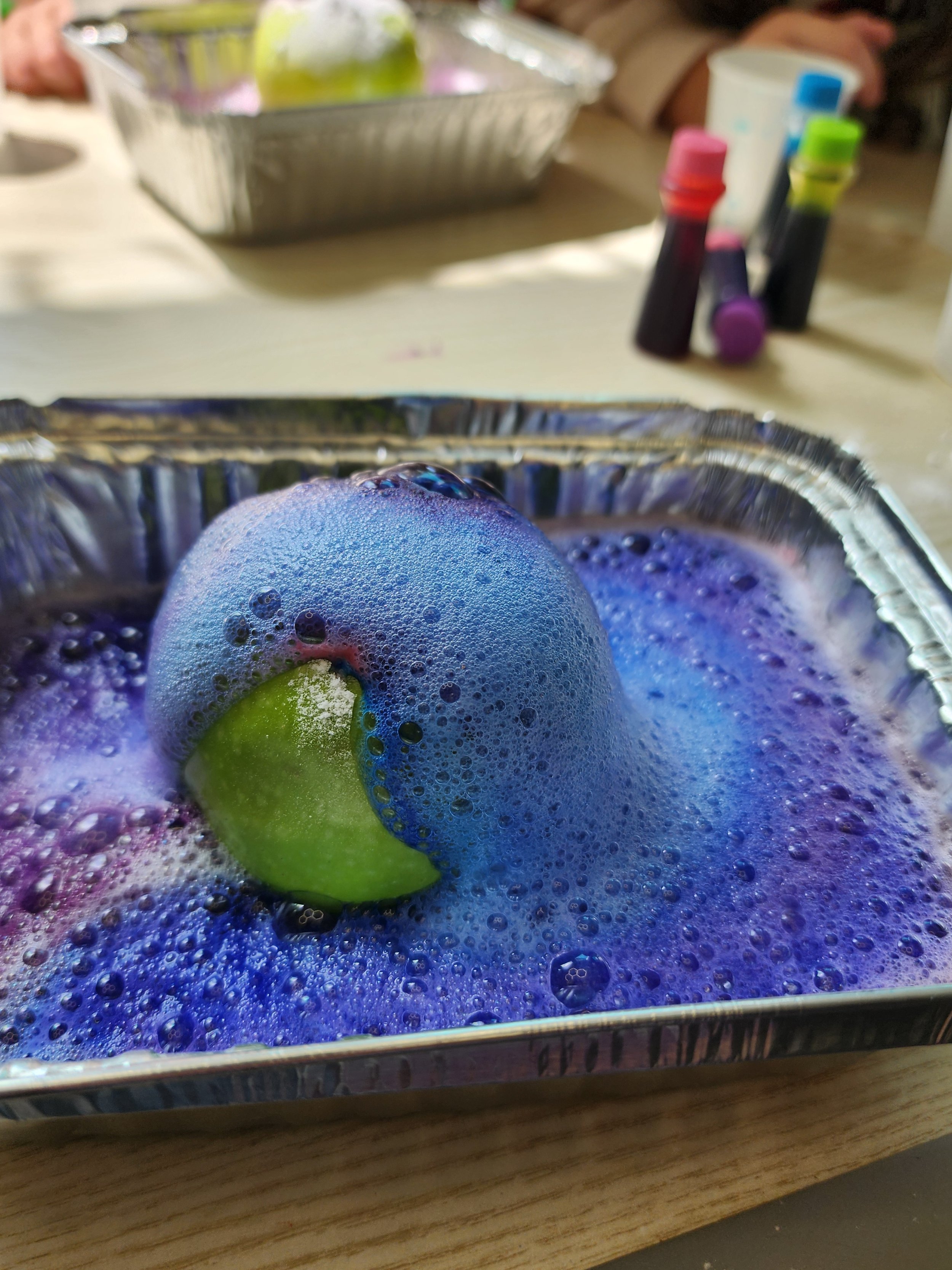 Foam on top of a green and purple ball inside a metal tray, with blue and purple coloring in the foam.