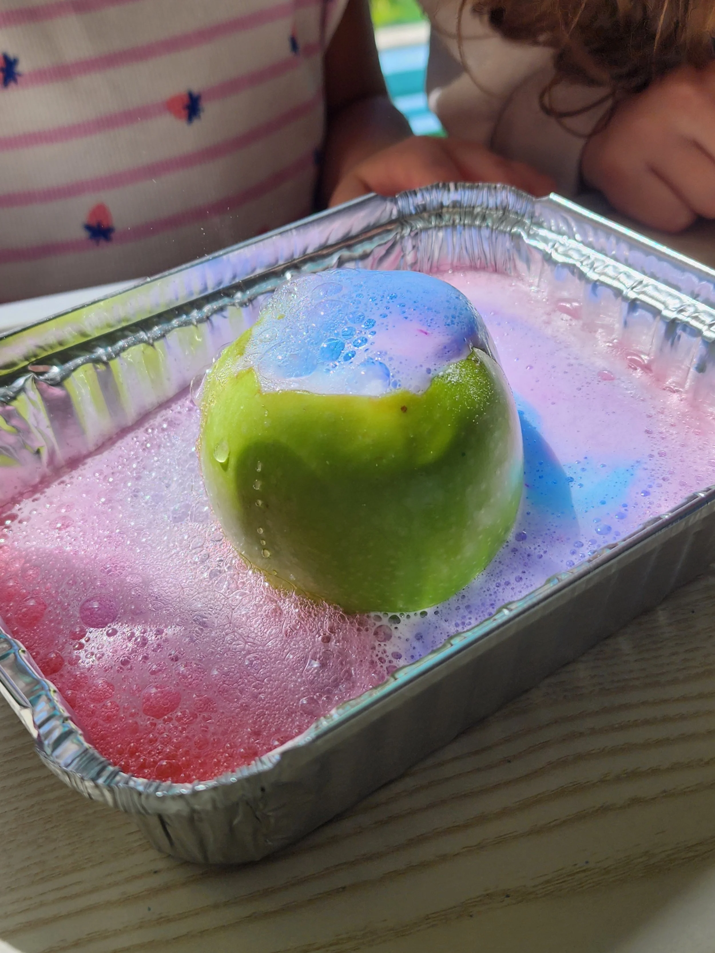 Green apple in a aluminum tray with colored water, soap and bubbles on it, children are watching it.