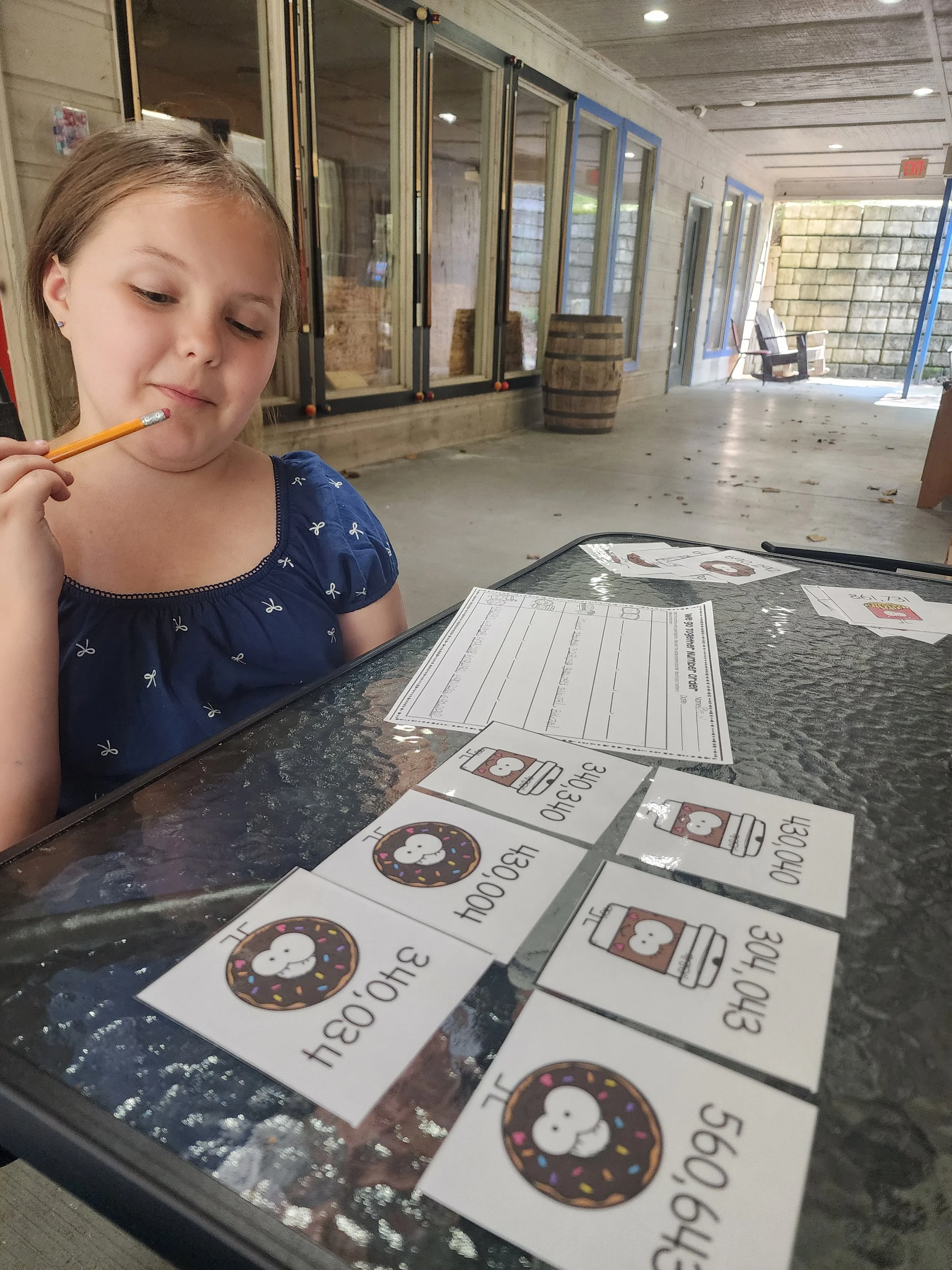 A young girl with long brown hair, wearing a blue shirt with small white bows, sits at an outdoor table looking at a worksheet and several flashcards with donuts, hot chocolate, and the word 'already' on them. The background shows a porch with glass doors and a stone wall.