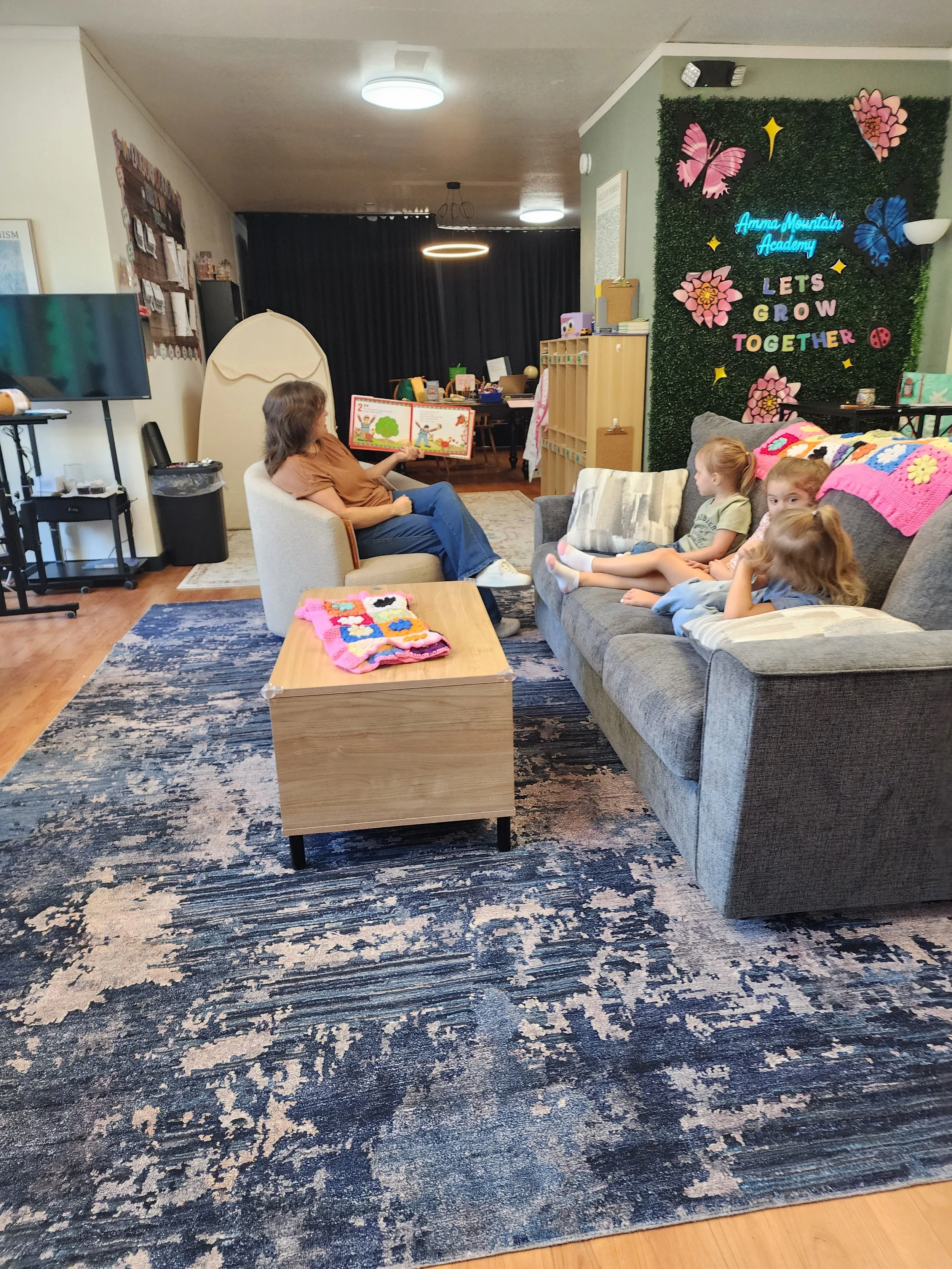 A woman reading a children's book to three young children in a cozy room with a navy blue patterned rug, wooden flooring, and a large gray sofa. The room has a decorative wall with butterflies and flowers, and a sign that says "Let's Grow Together."
