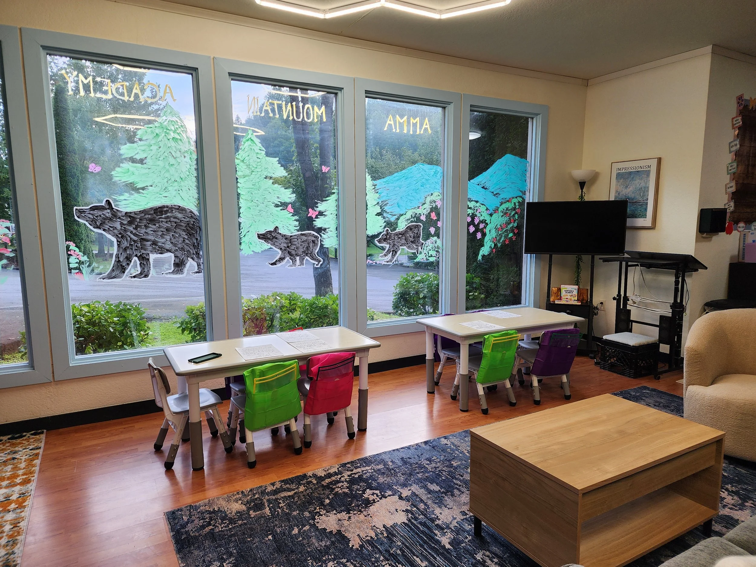 Classroom window with colorful animal and nature-themed decorations painted on the glass, including bears, trees, and butterflies, with a view of outdoors and a classroom interior featuring small chairs, tables, a television, and framed artwork.