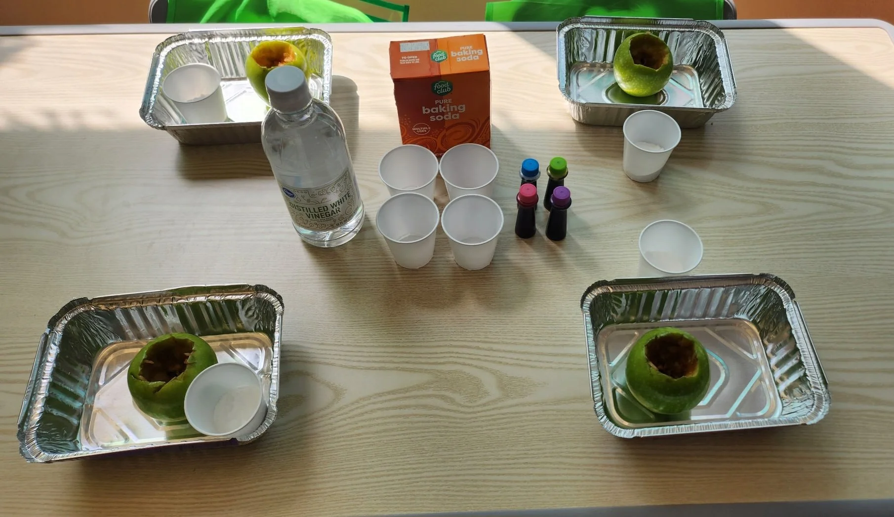 Four aluminum trays each containing a hollowed green apple, with a white cup nearby. In the center, there are four small cups, a bottle of distilled white vinegar, a box of baking soda, and four small bottles of liquid dye.