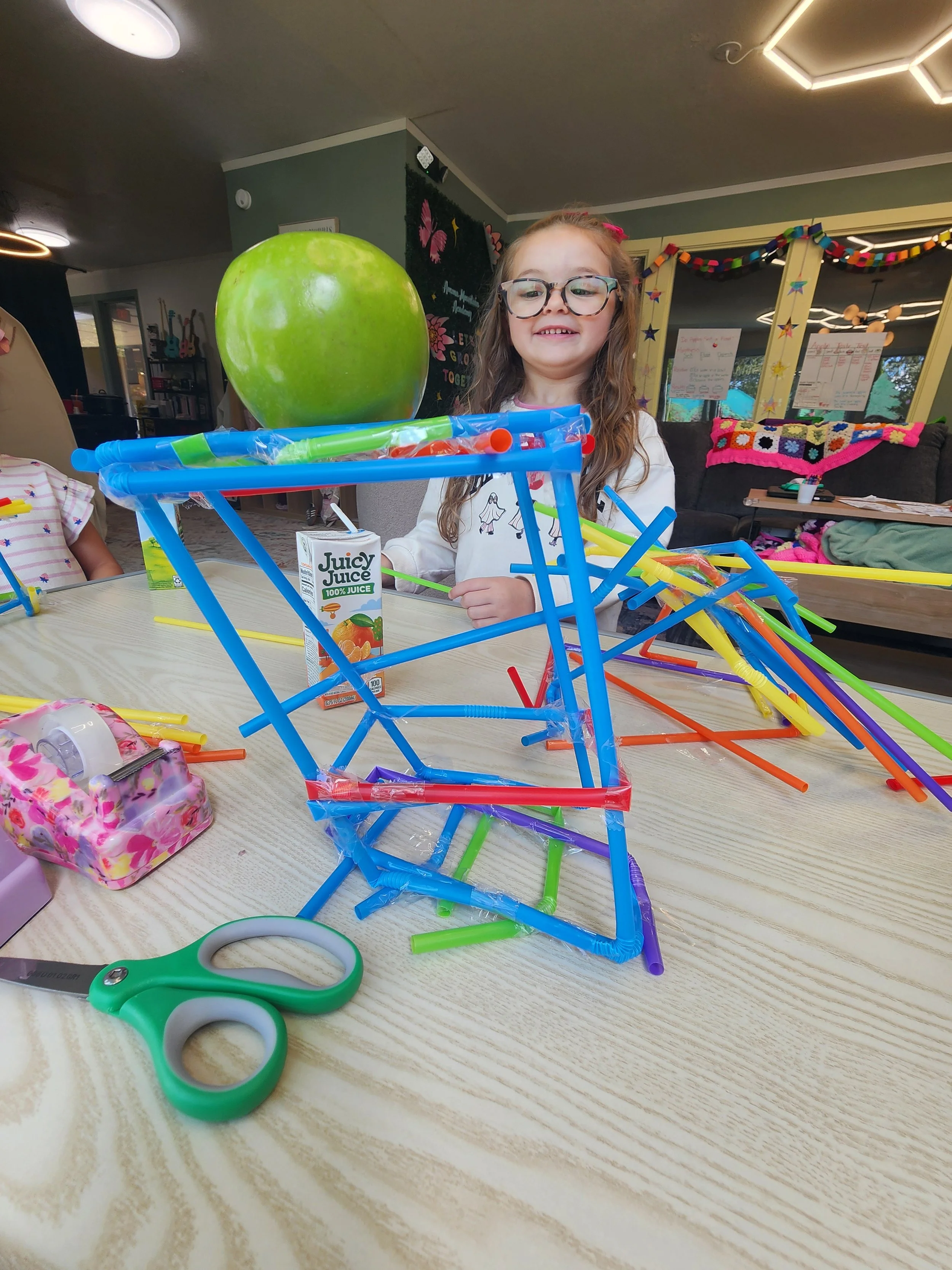 A young girl with glasses smiling at a table filled with colorful plastic straws, a small juice box, and a pair of green scissors. In the background, a green apple, a decorated chalkboard, and colorful paper garlands are visible.
