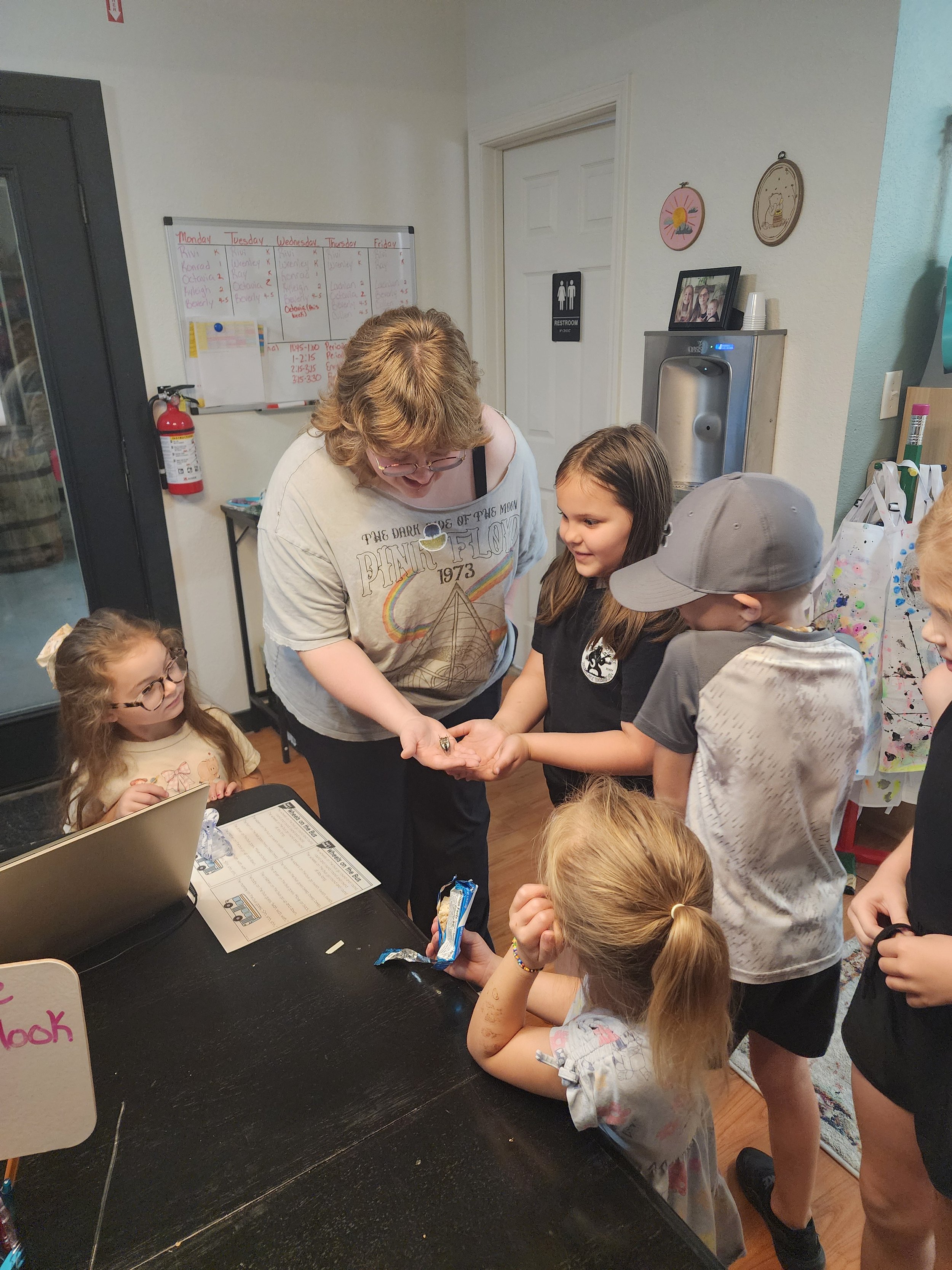 A group of children and a woman are gathered around a table with a laptop and a paper, engaging in an activity. The room has posters, a water fountain, and a whiteboard with a weekly schedule on the wall.