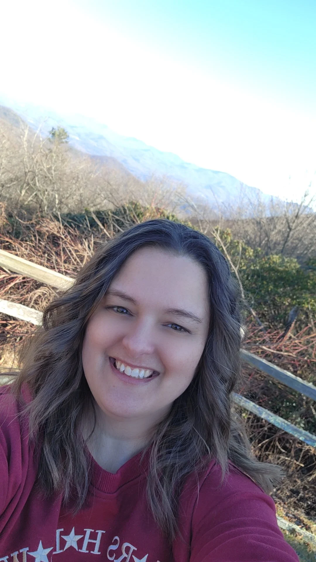 A woman with shoulder-length wavy hair and fair skin smiling at the camera outdoors with mountains, trees, and a clear sky in the background.