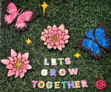 Decorative wall with pink and blue butterflies, pink flowers, yellow stars, and colorful letters spelling 'LET'S GROW TOGETHER' on a green hedge background.