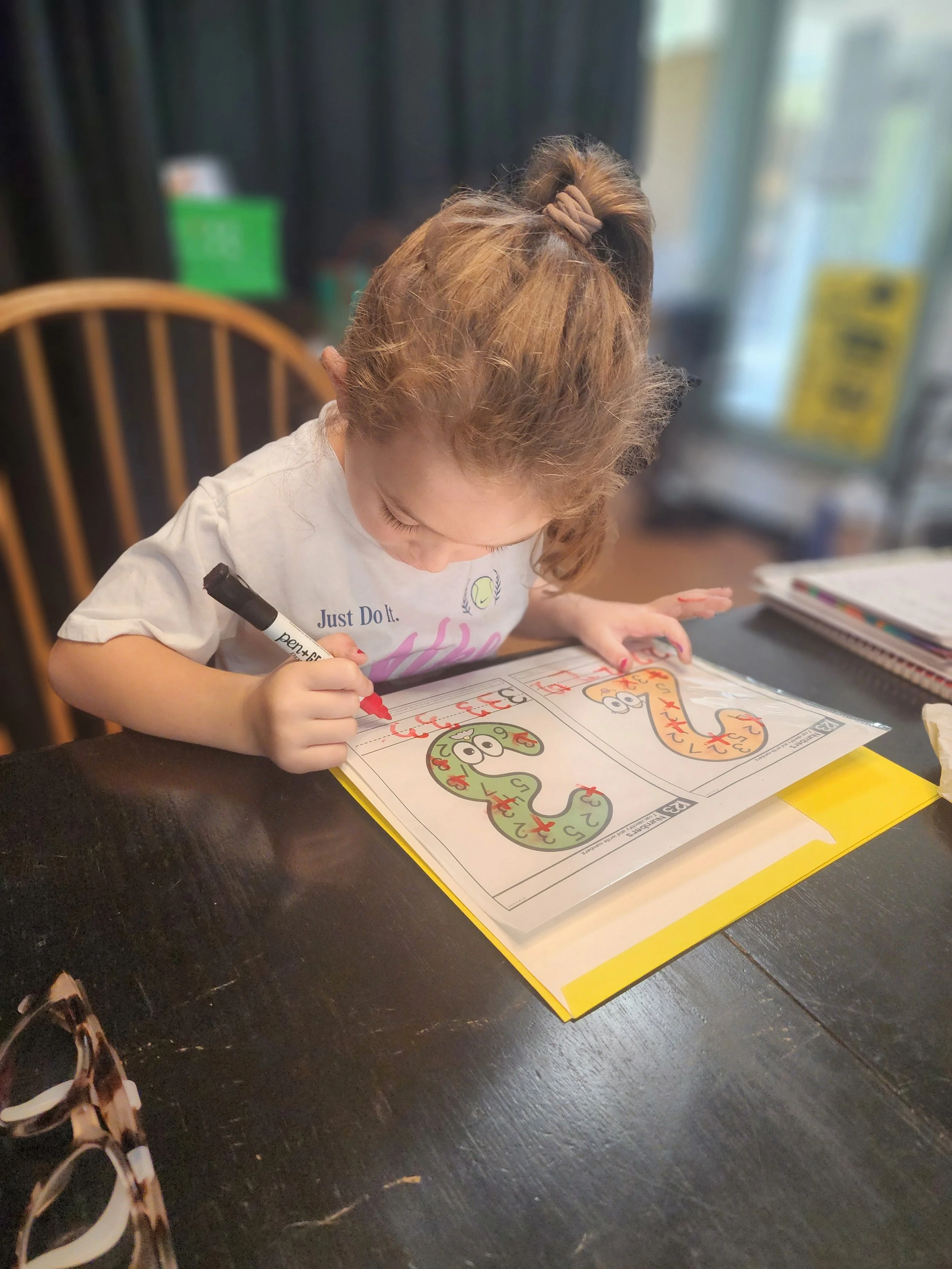 A young girl with curly hair in a ponytail is coloring on a worksheet with a red marker. The worksheet features illustrations of cartoonish snakes with numbered sections, and the girl is focused on coloring the snake's tongue red. She is sitting at a dark wooden table, and there are notebooks and a bag on her right side.