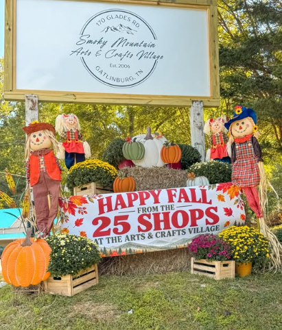 Fall display with scarecrows, pumpkins, and mum plants at a craft village, with a sign thanking 25 shops for the fall celebration.