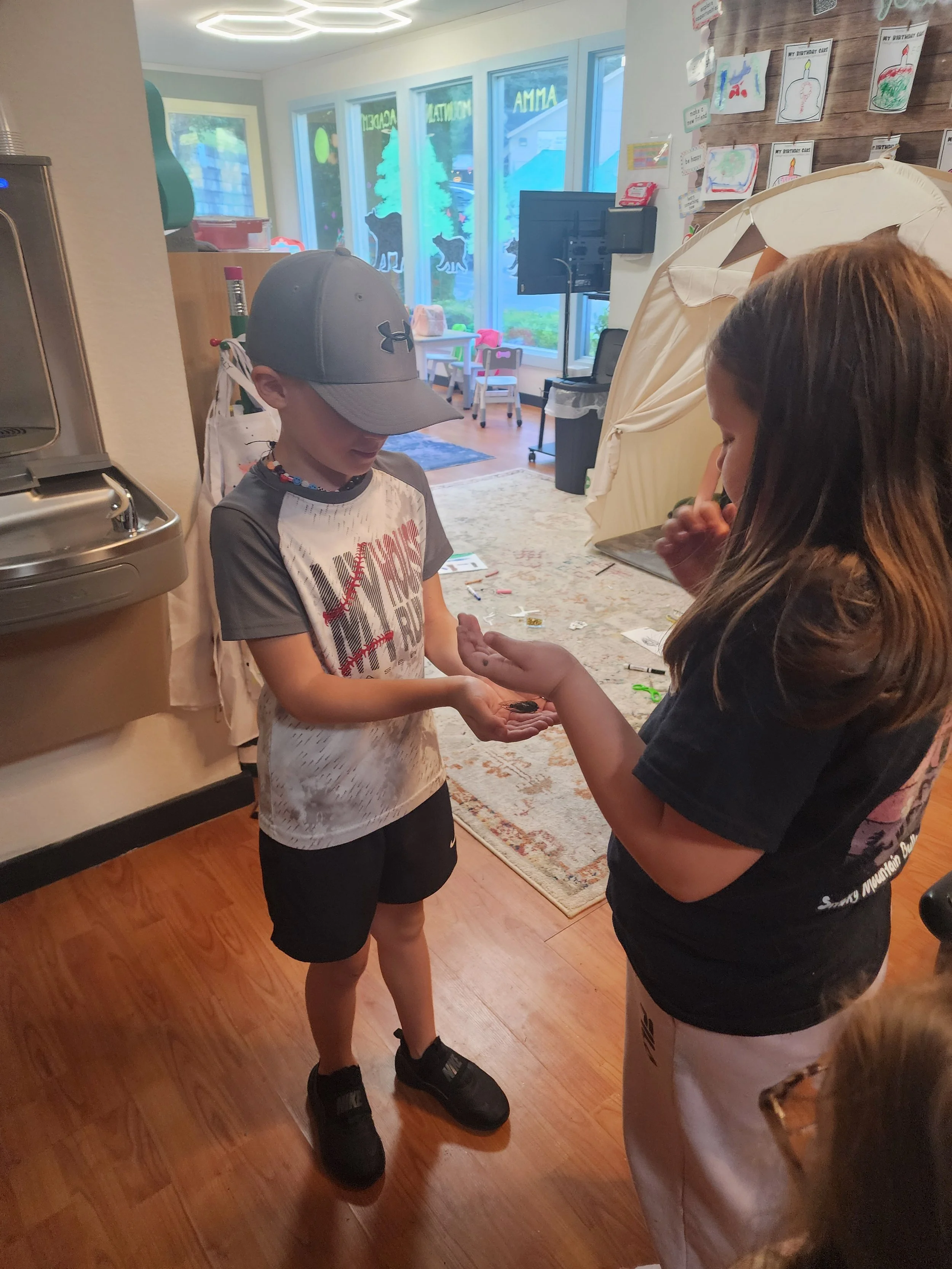 A boy and a woman exchanging a coin inside a room with a rug, a wall decorated with children's drawings, and a window with Christmas-themed window decals.