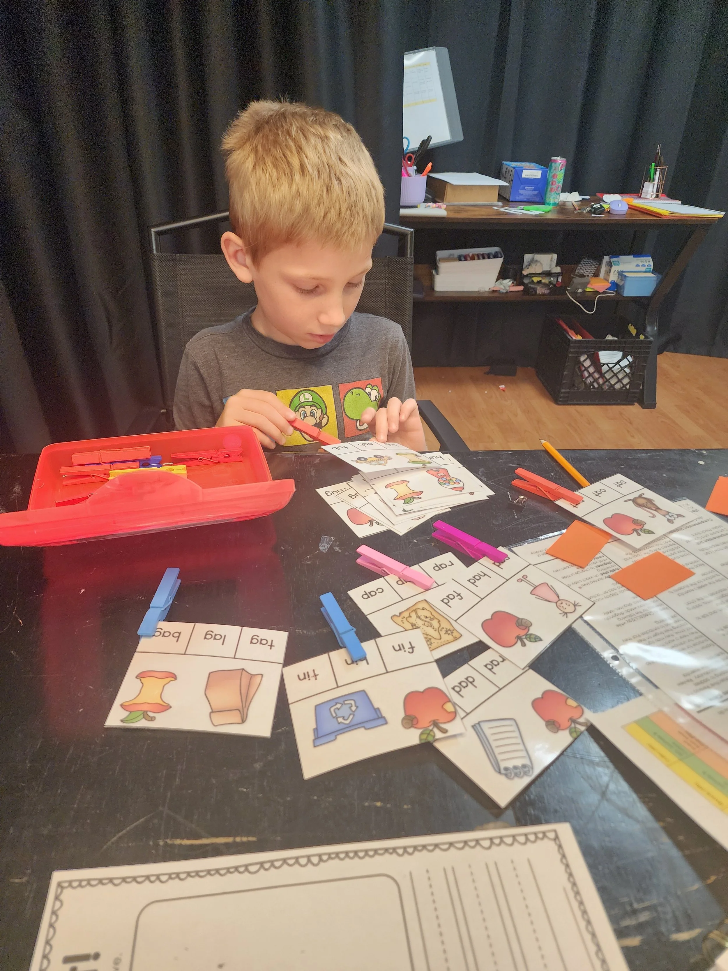 A young boy with short blond hair engaged in a reading activity with picture cards, sitting at a dark table with a red plastic container of clothespins and various educational materials and supplies scattered around. In the background, a black curtain and a wooden desk with office supplies and containers are visible.