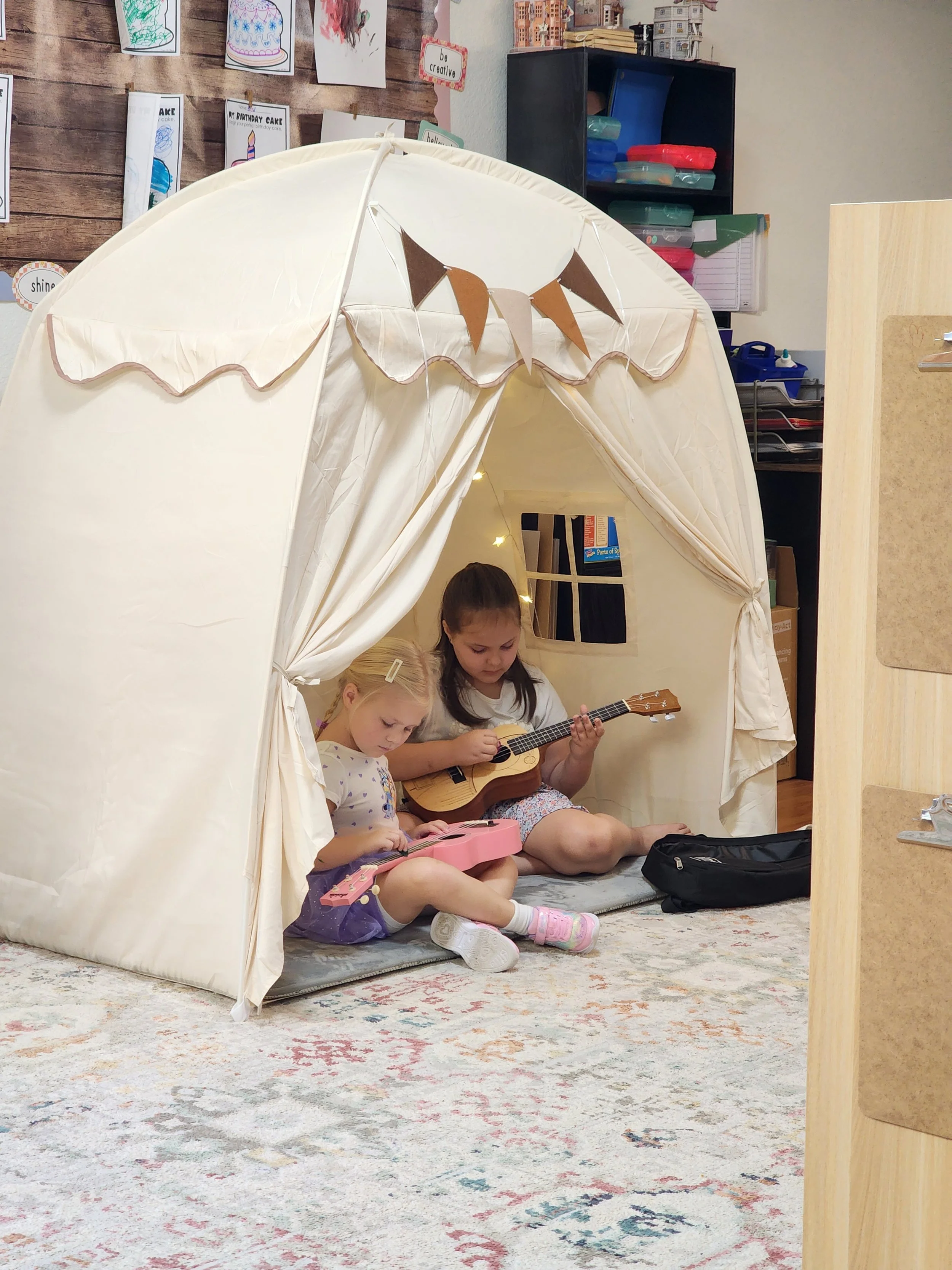 Two children sitting in a cozy indoor play tent, playing guitars. The girl on the left has blonde hair, and the girl on the right has dark hair. The tent is cream-colored with bunting decorations on the top. The room has a colorful patterned rug and shelves filled with storage boxes in the background.