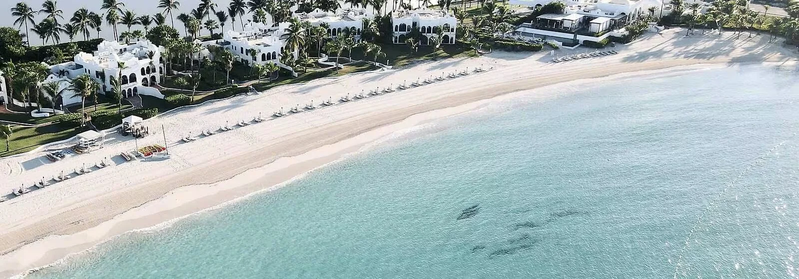Aerial view of a beach with white sand, turquoise water, lined with lounge chairs and umbrellas, and a row of white buildings with palm trees behind them.