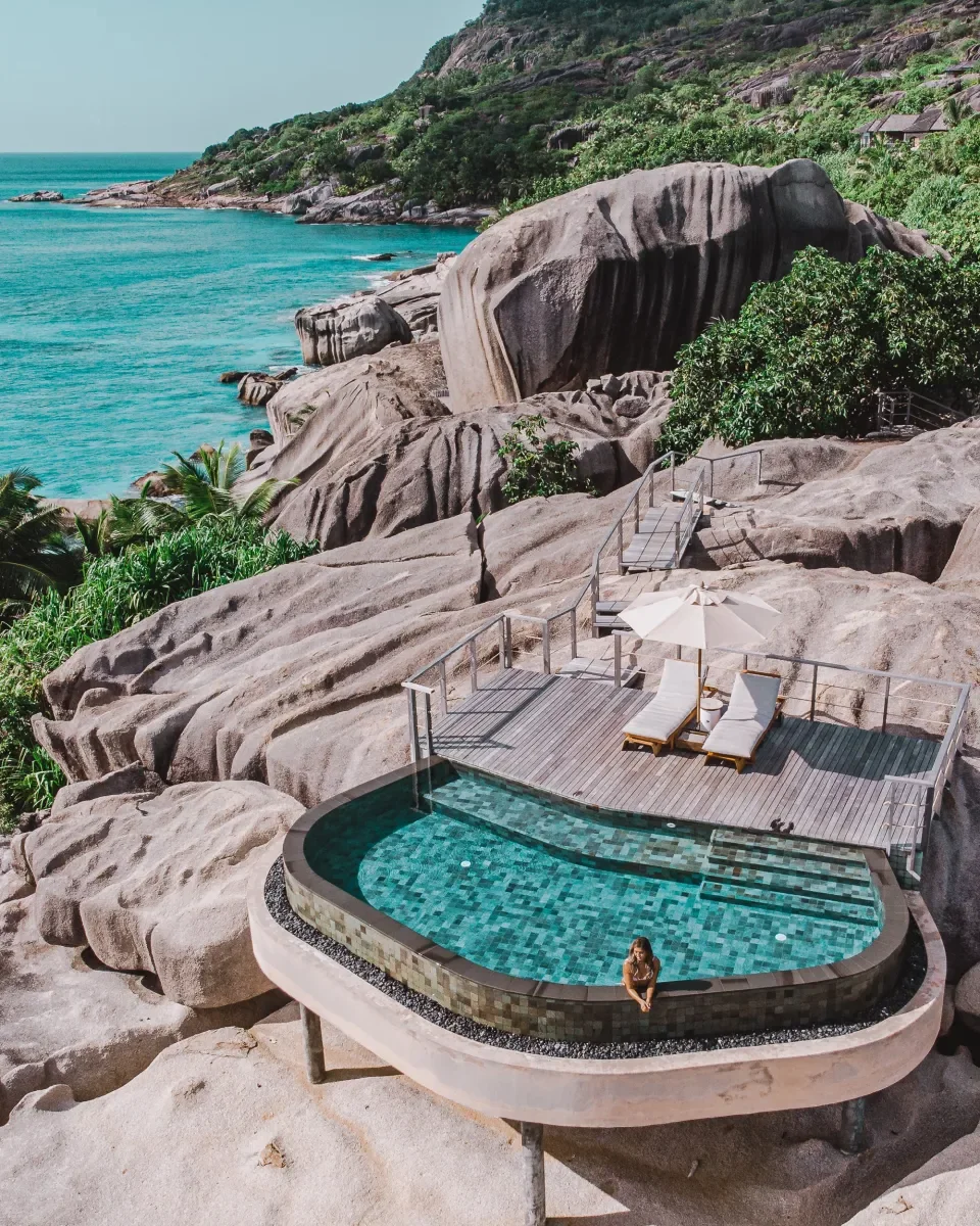 A rooftop pool on rocky terrain overlooking a tropical ocean with green hills, a deck with lounge chairs and an umbrella.