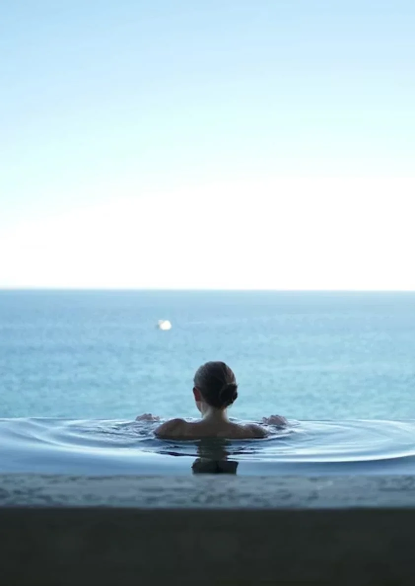 A person with hair tied back in a bun is relaxing in an infinity pool, looking out over the ocean and blue sky.