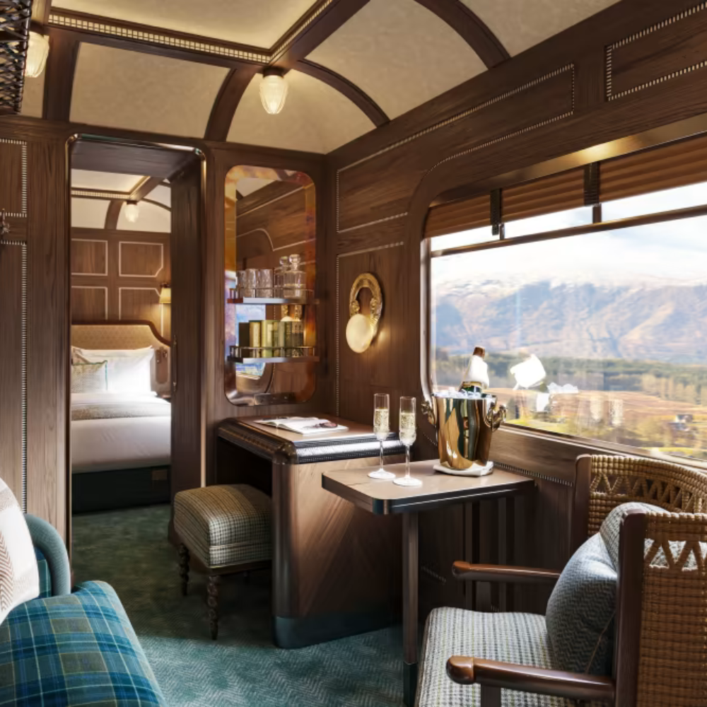 A luxurious train cabin with wood-paneled walls, a window overlooking mountains, a small table with champagne and glasses, a cozy armchair, and a separate sleeping area with a bed.