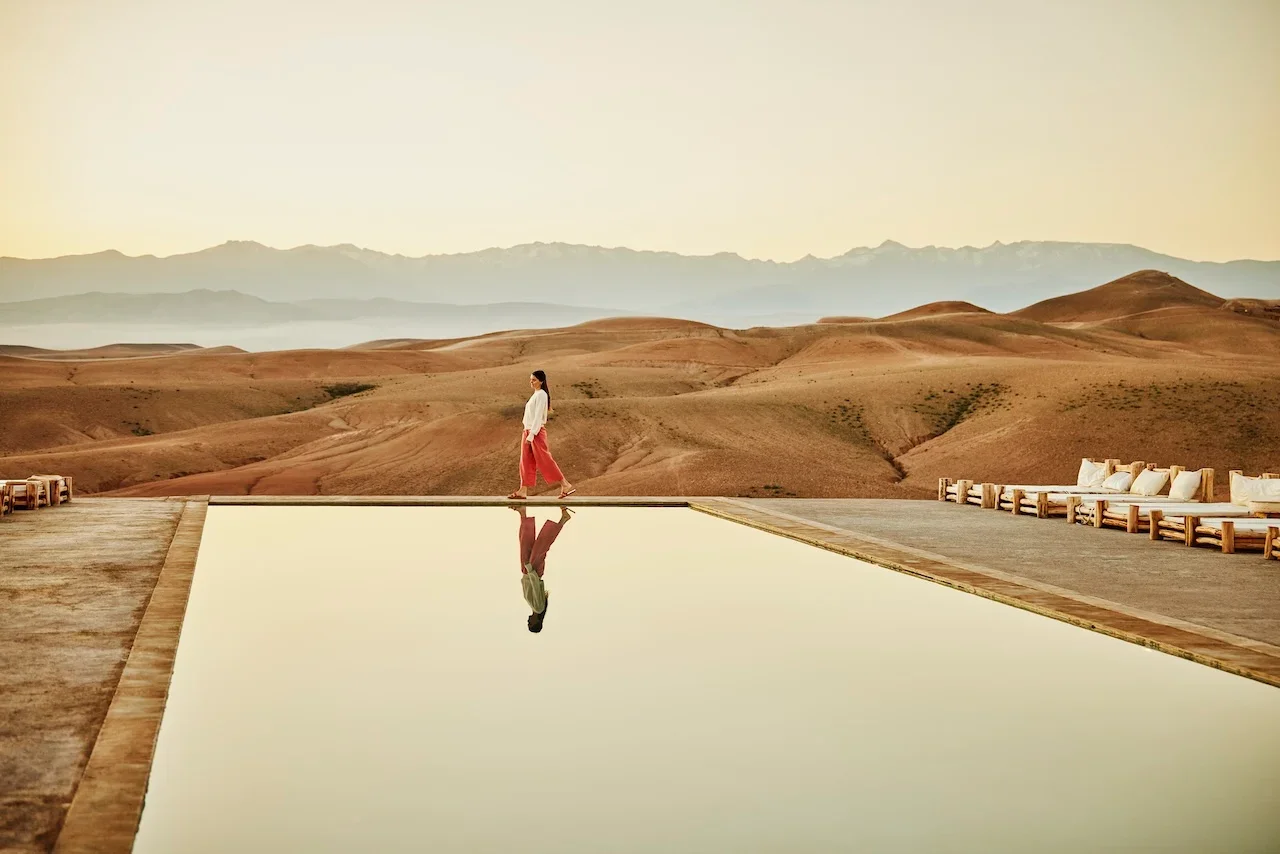 A woman walks along the edge of an infinity pool with a desert landscape and mountains in the background during sunset or sunrise.