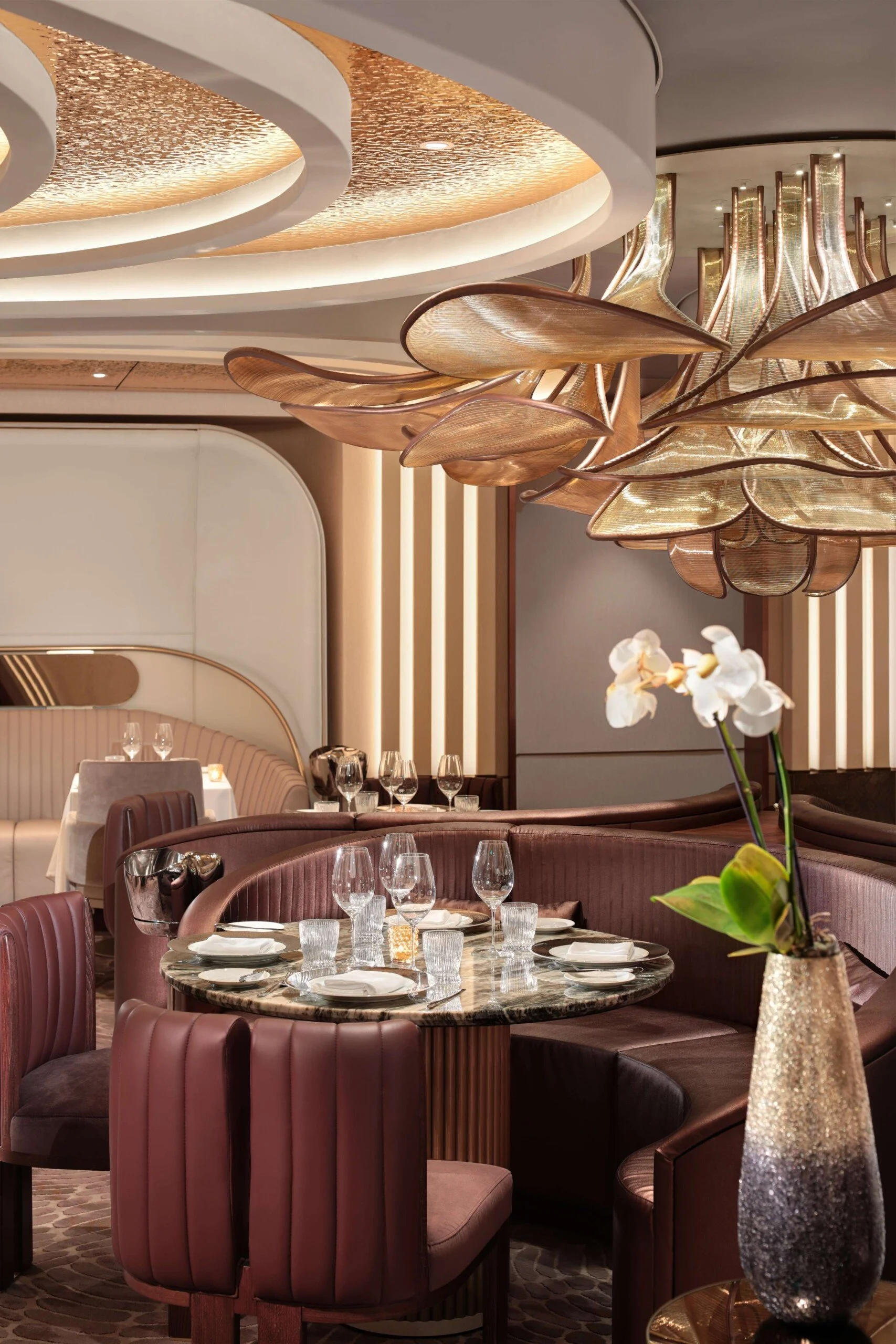A luxurious restaurant interior with a round table set with wine glasses, plates, and cutlery, surrounded by plush, pink upholstered chairs and a curved banquette. Ornamental lighting fixtures hang from the ceiling, and a tall vase with white orchids decorates the table.