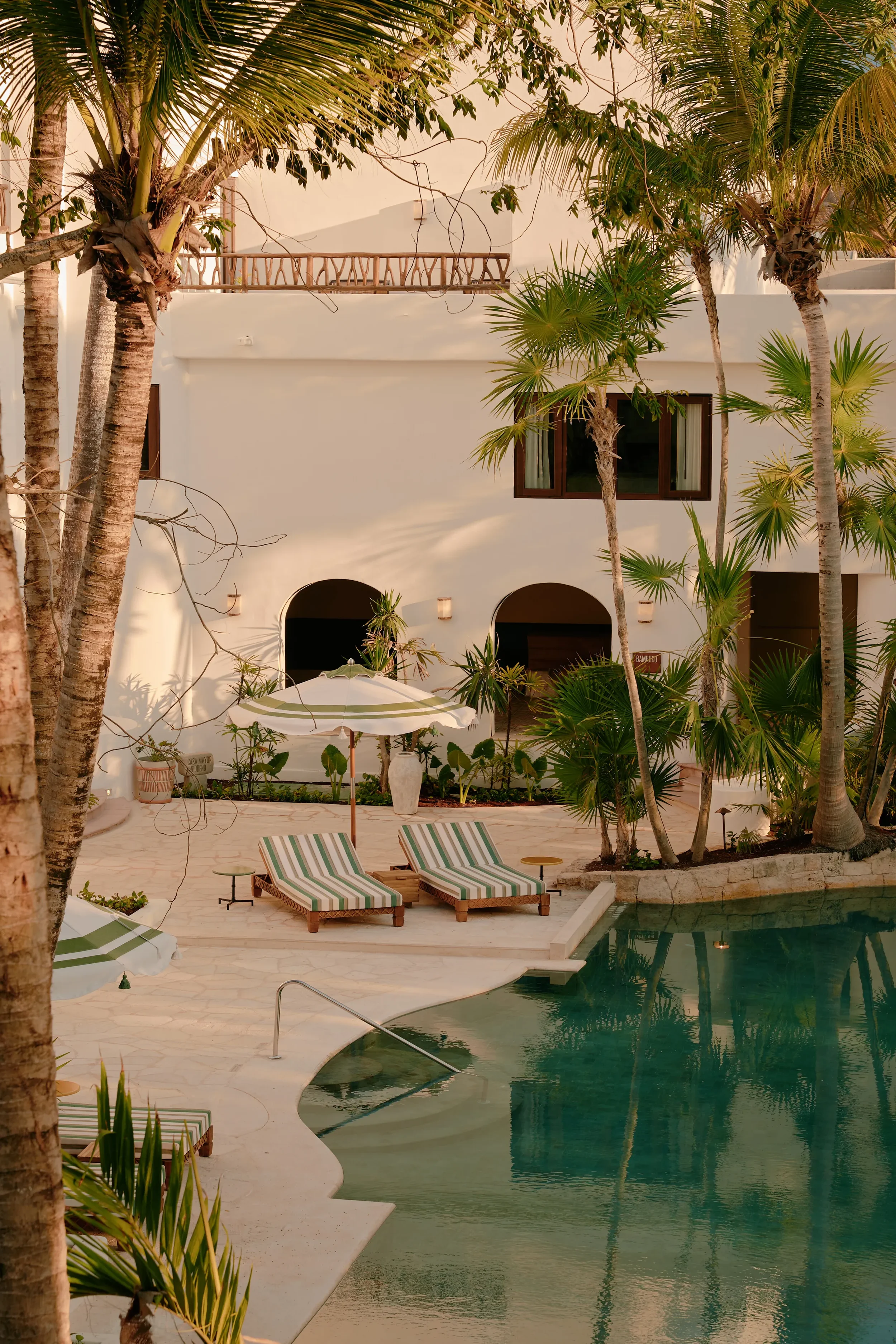 View of a swimming pool surrounded by palm trees, lounge chairs with striped cushions, and a white building with arched doorways and windows.