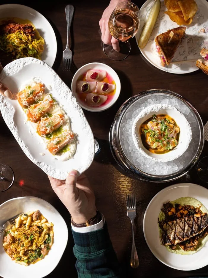 Various dishes on a dinner table, including sushi, seafood, salad, and other appetizers, with glasses of wine.