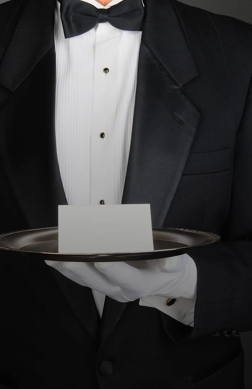 A waiter in a tuxedo holding a tray with a blank white card, wearing white gloves.