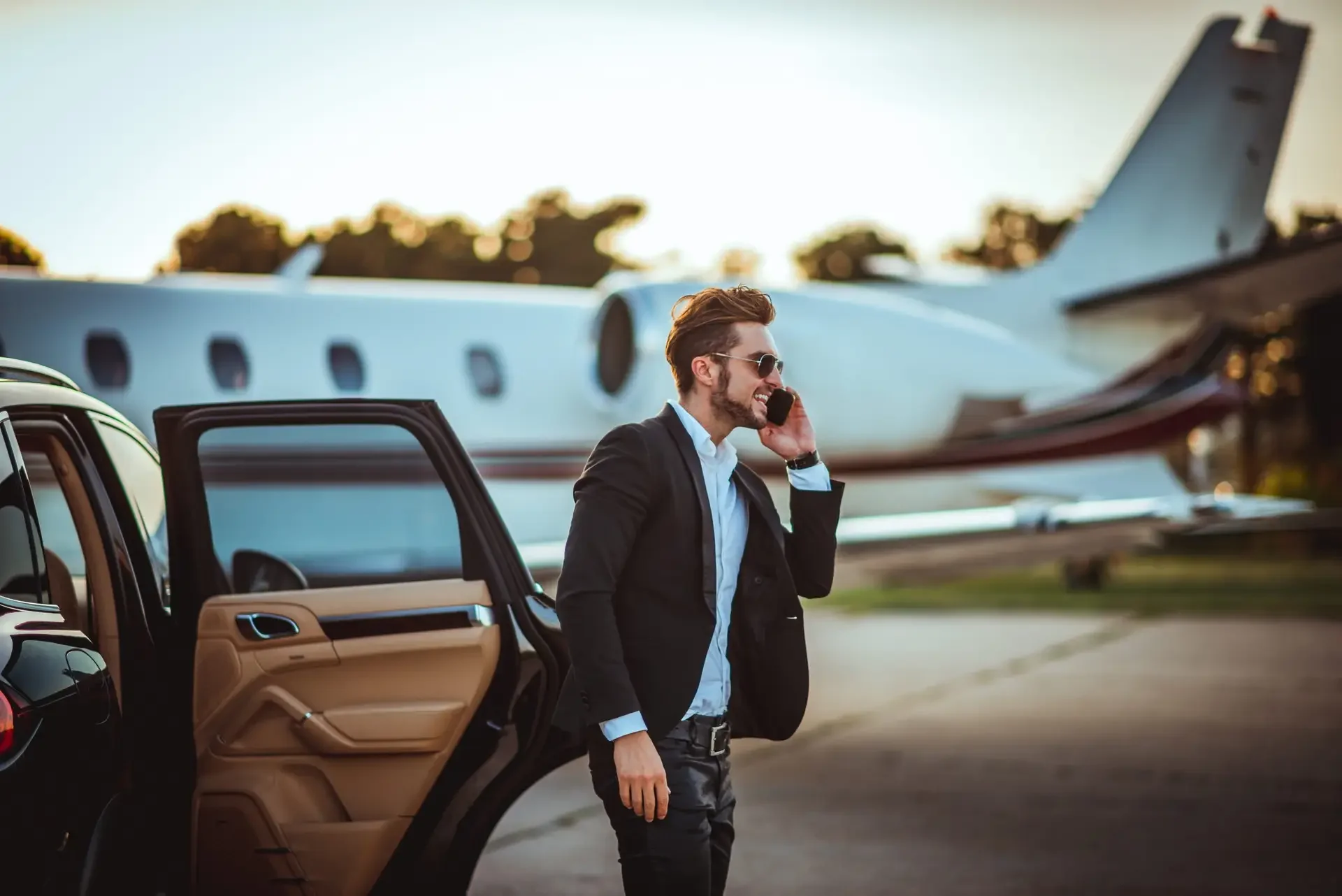 A man in a black suit and sunglasses standing near a luxury car, talking on his phone, with a private jet in the background, during sunset.