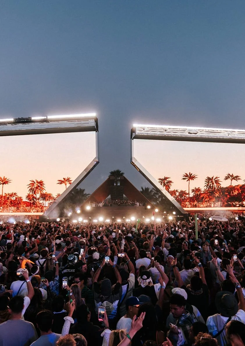 Crowd at an outdoor concert with stage and palm trees in background during sunset.