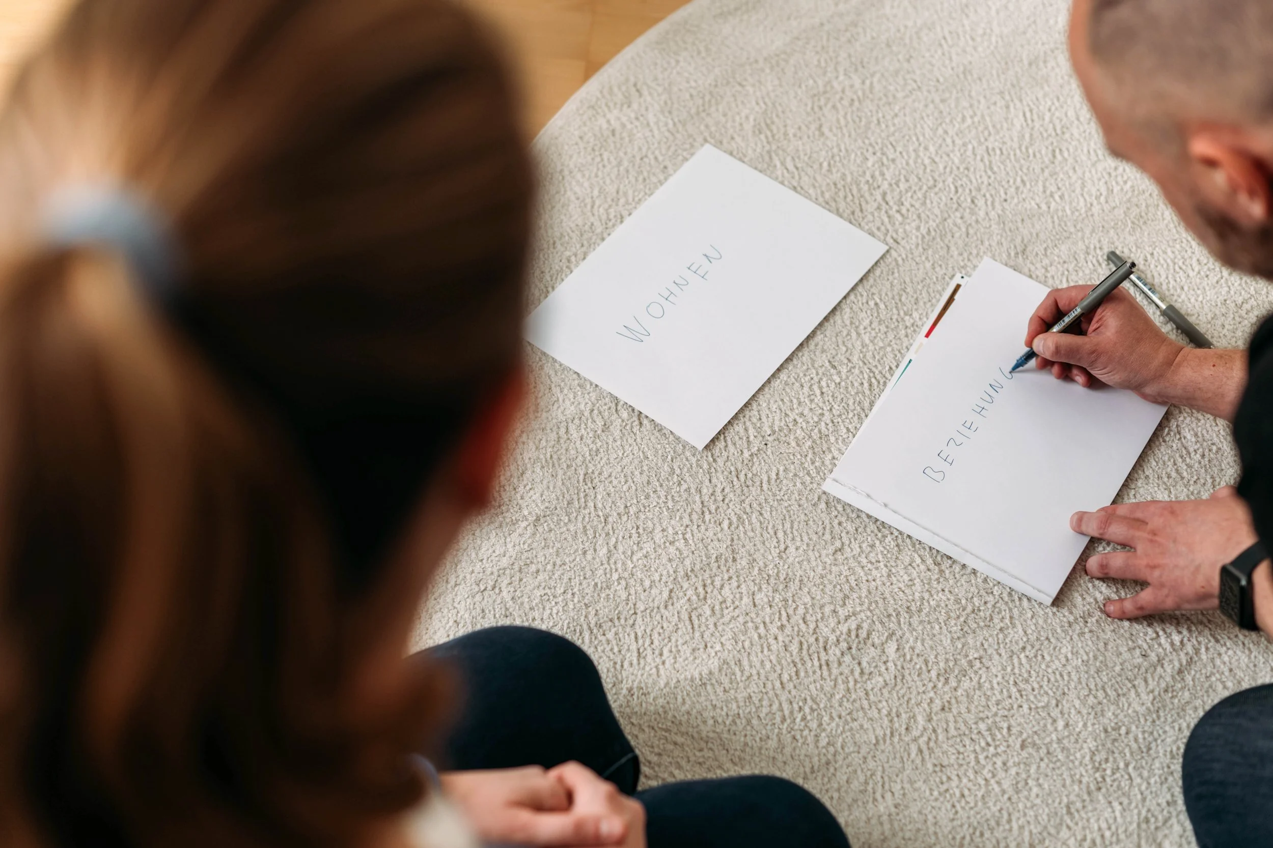Zwei Personen unterhalten sich, während einer der beiden auf dem Boden sitzt und auf eine weiße Karte schreibt. Die Karte hat das Wort 'Beziehung' darauf, die andere Karte hat 'Wohnen' darauf.