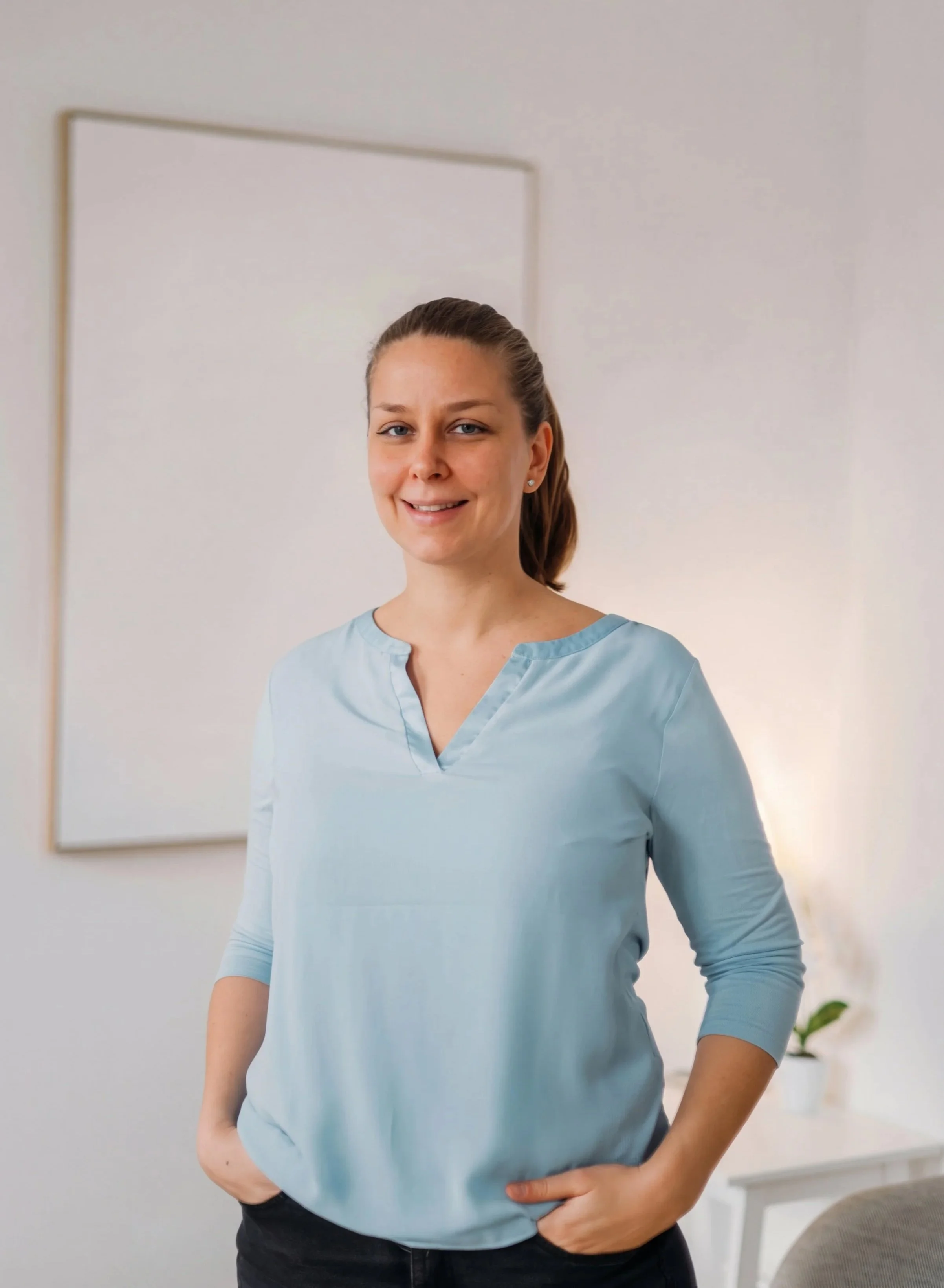 Eine lächelnde Frau mit braunem, mittellangem Pferdeschwanz steht vor einem weißen Hintergrund, trägt eine hellblaue Bluse und hat die Hände in den Taschen.