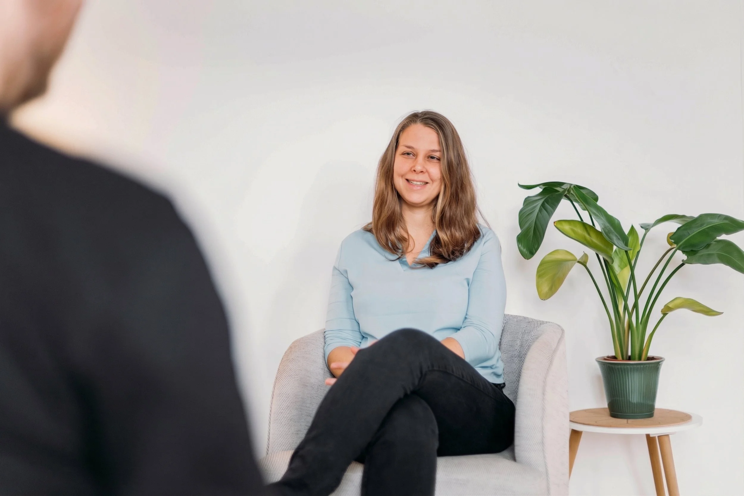 Frau im Gespräch, sitzend auf einem Stuhl mit einer Pflanzen im Hintergrund.