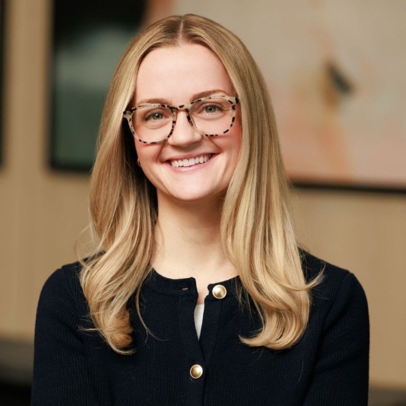 Smiling woman with long blonde hair and glasses, wearing a black top with gold buttons, indoors with a blurred background.
