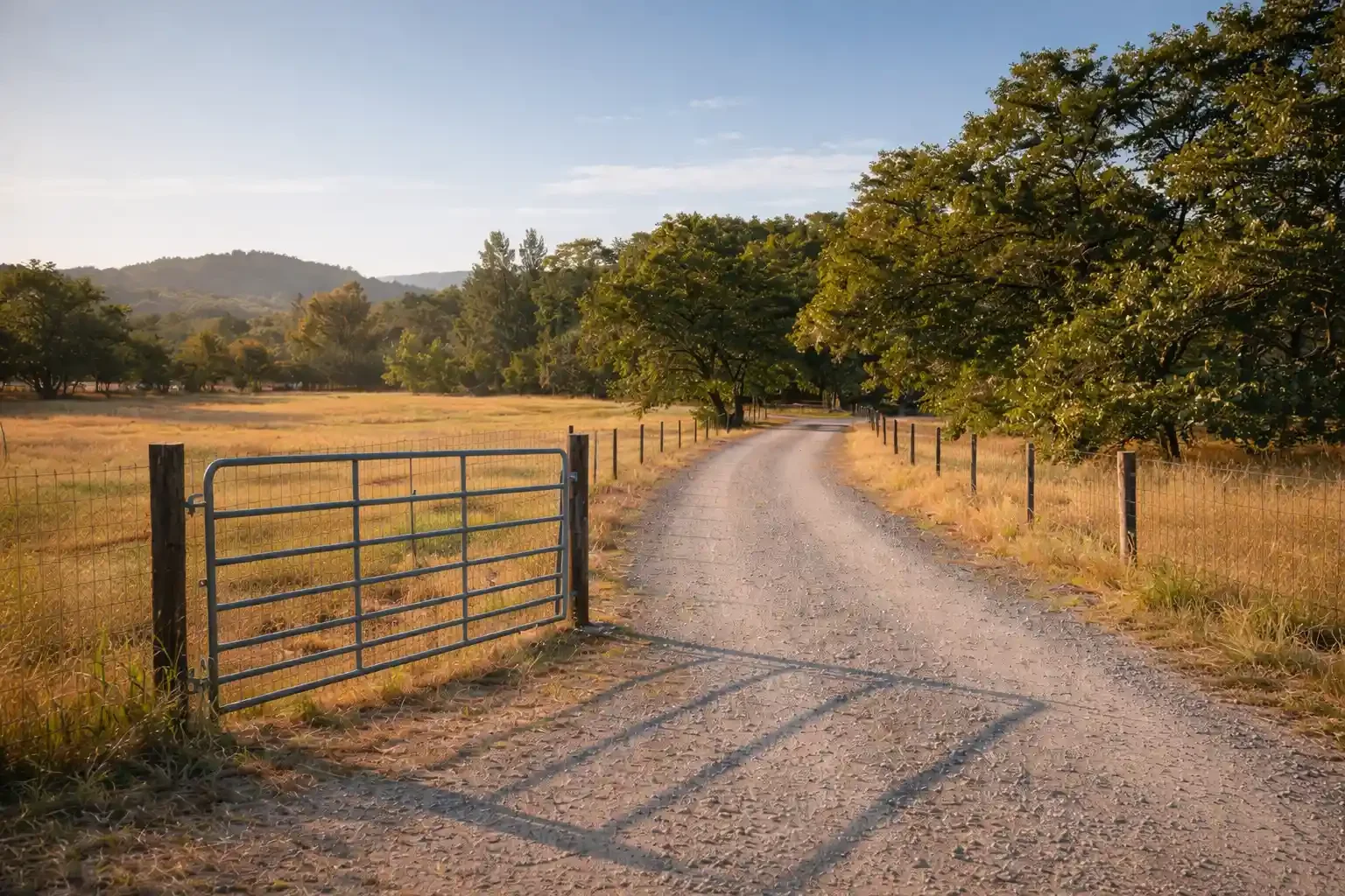 A dirt country road with a metal gate on the left and fencing on both sides, surrounded by trees and open fields during daytime.