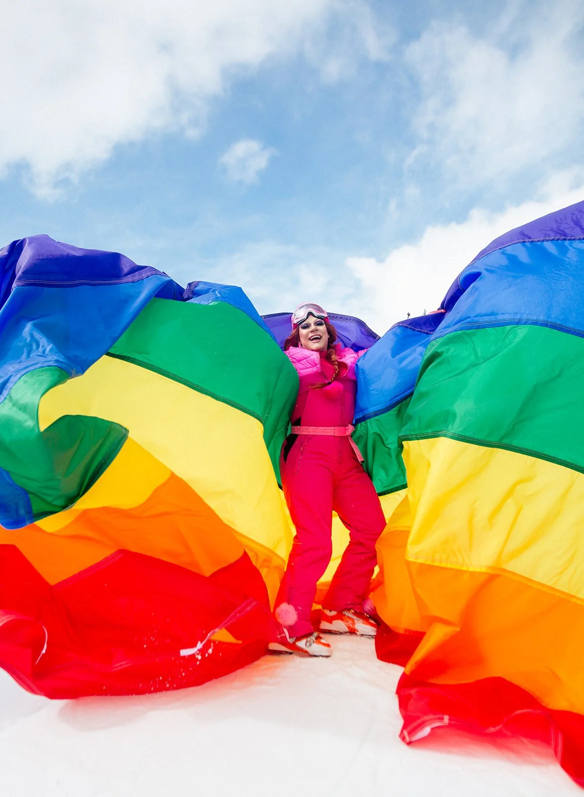 A woman in ski gear, including a white helmet, goggles, a teal jacket, and pink skis, is joyfully in powder snow in a snowy forest with tall snow-covered trees and a clear blue sky.