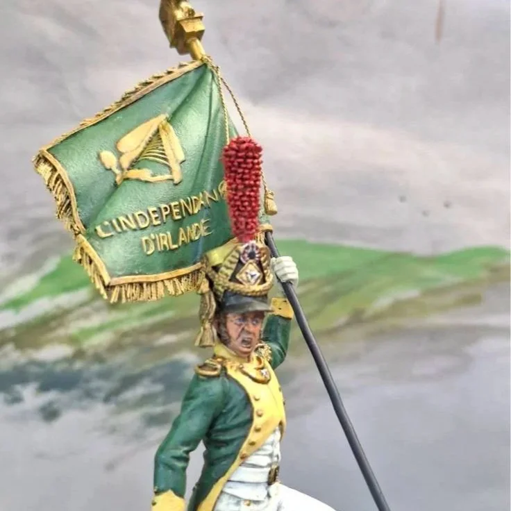 Miniature toy soldier dressed in green and yellow uniform, holding a flag that reads 'Lindepedance D'lan' with a hill and cloudy sky background.