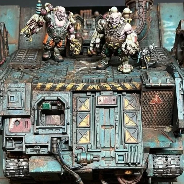 Miniature figures of two large, armored alien-like creatures with weapons, standing on a detailed sci-fi or industrial-themed diorama or model set. The scene features a weathered, painted surface with intricate machinery and metallic textures.