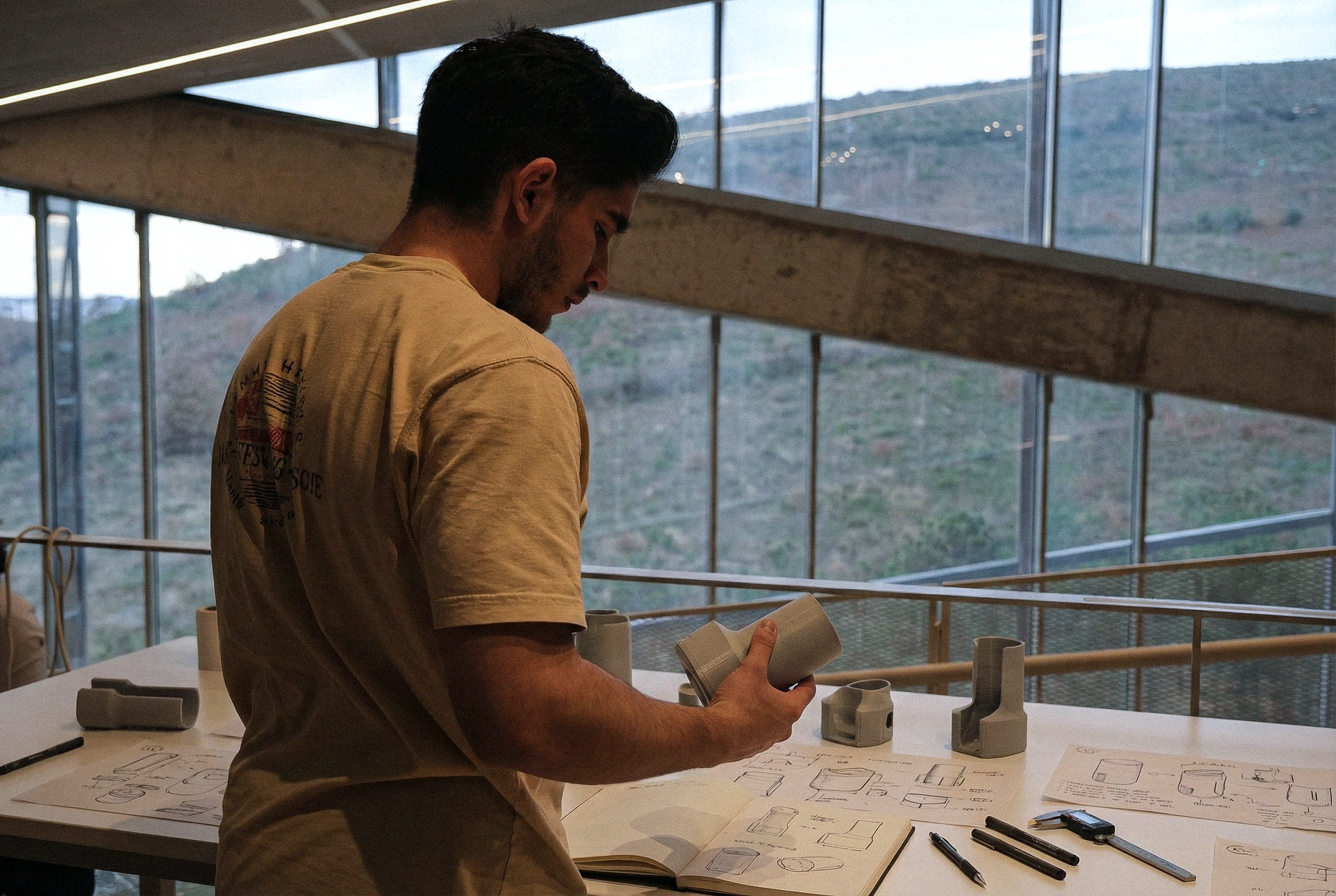 A man in a tan T-shirt examining a ceramic vessel with sketches and design plans on a table in front of him. Large windows with a view of a hillside are in the background.
