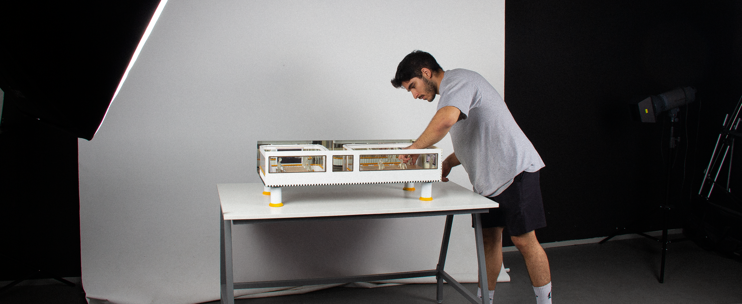A young man in a gray T-shirt and black shorts working on a small architectural model or experiment on a white table in a professional photo studio with black curtains and studio lights.
