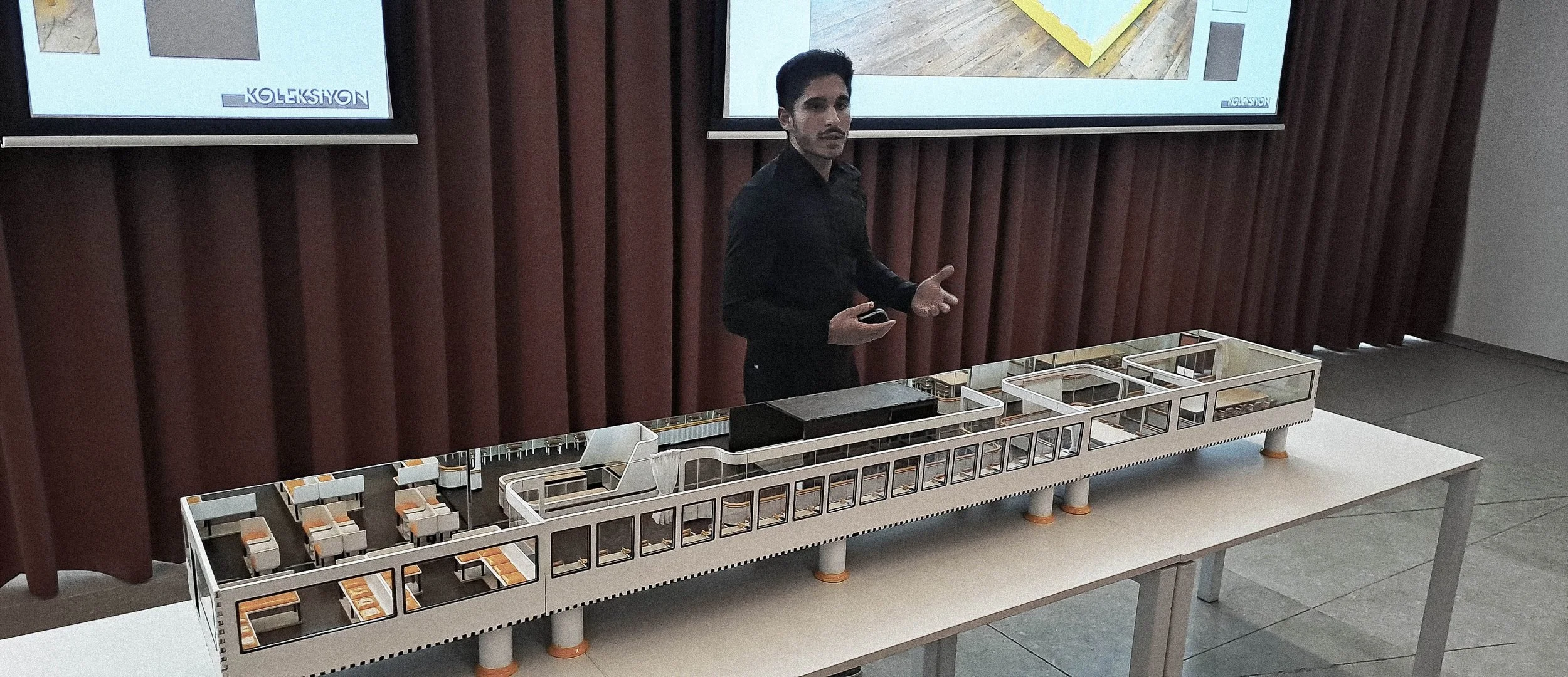 A man in black clothing presenting a scale model of a building or structure at a table, with two large screens displaying images in the background.