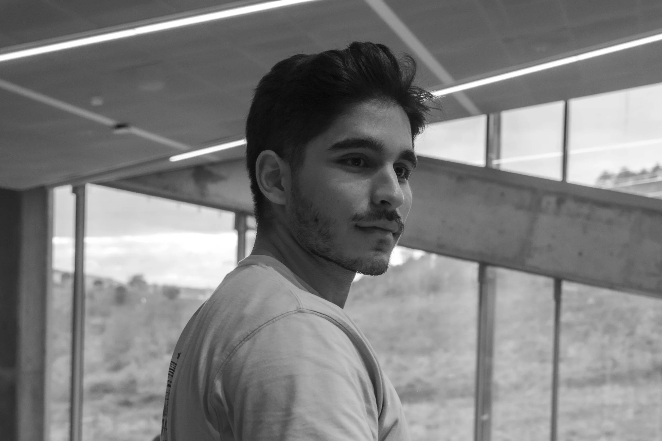 A young man with dark hair and a beard looking over his shoulder in a building with large windows and a scenic landscape outside.