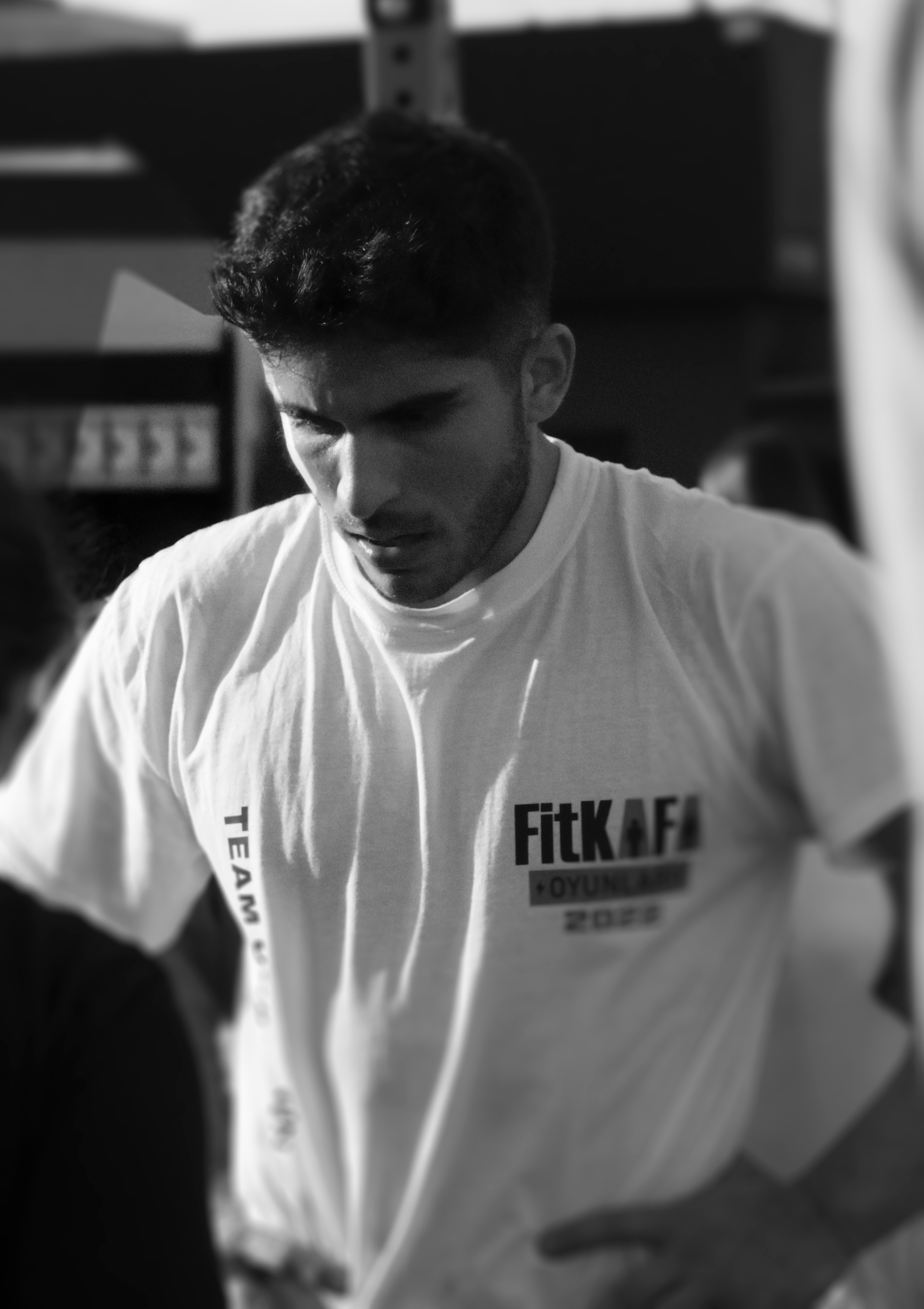 A young man with dark hair and a beard, wearing a white T-shirt with the words "FitKAFE" and other text, looking down with a focused expression.