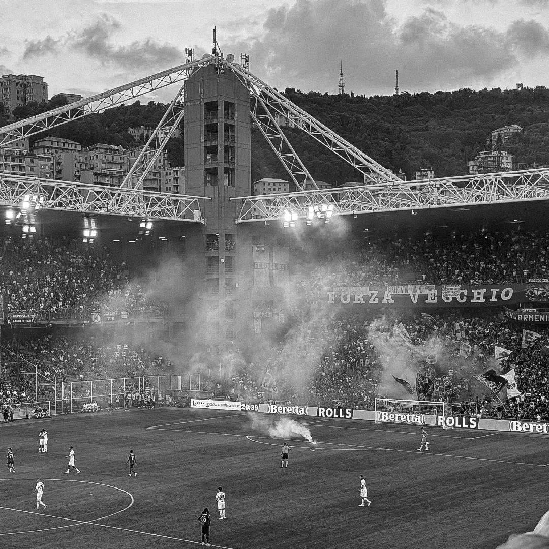 A black and white photo of a soccer stadium filled with spectators, with banners and flags, some smoke from flares, and a few players on the field. Hills and buildings are visible in the background.