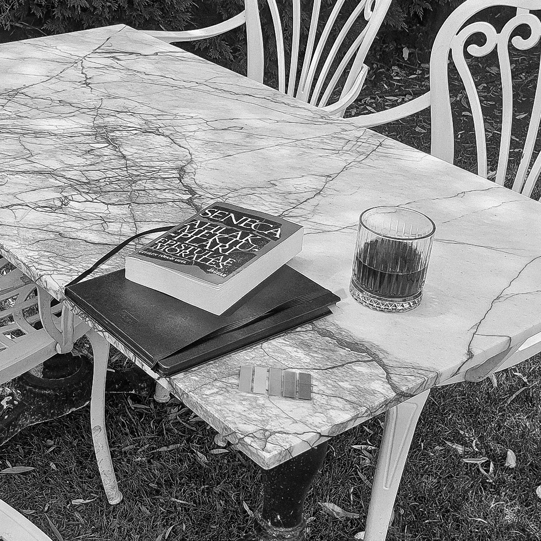A marble outdoor table with a glass of dark beverage, a stack of books, a black notebook, a color swatch, and a paperback with a cover featuring text, set in a garden with white metal chairs in the background.