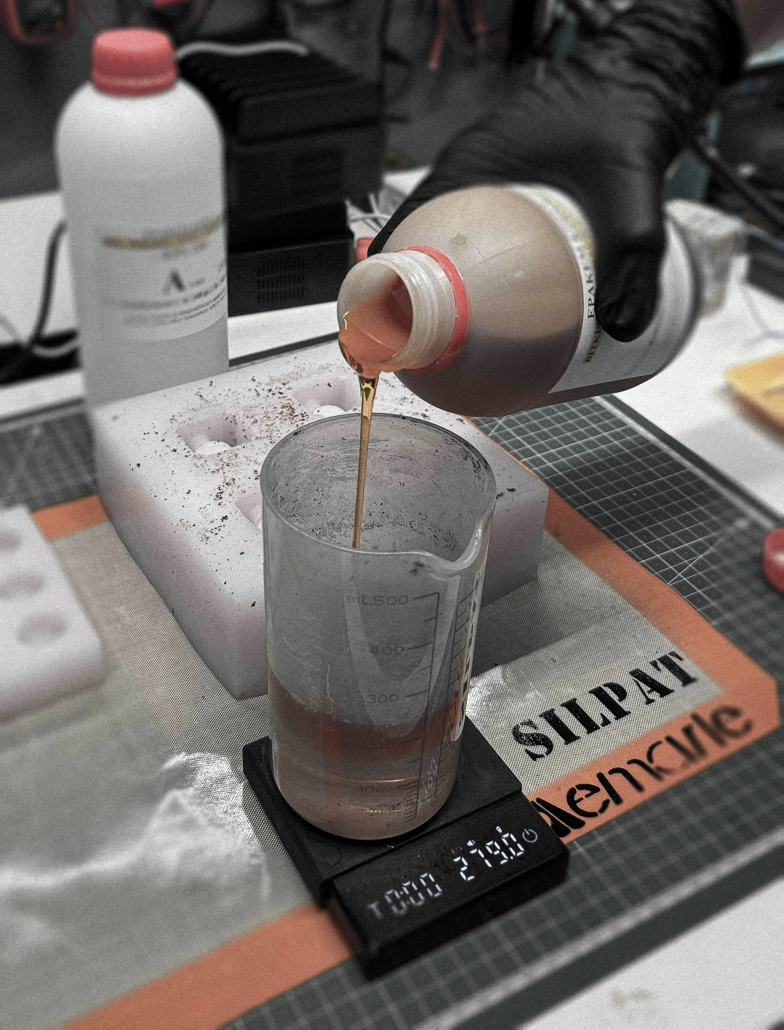 A person wearing black gloves is pouring a reddish-brown liquid from a bottle into a beaker on a digital scale, with various laboratory supplies and equipment in the background.