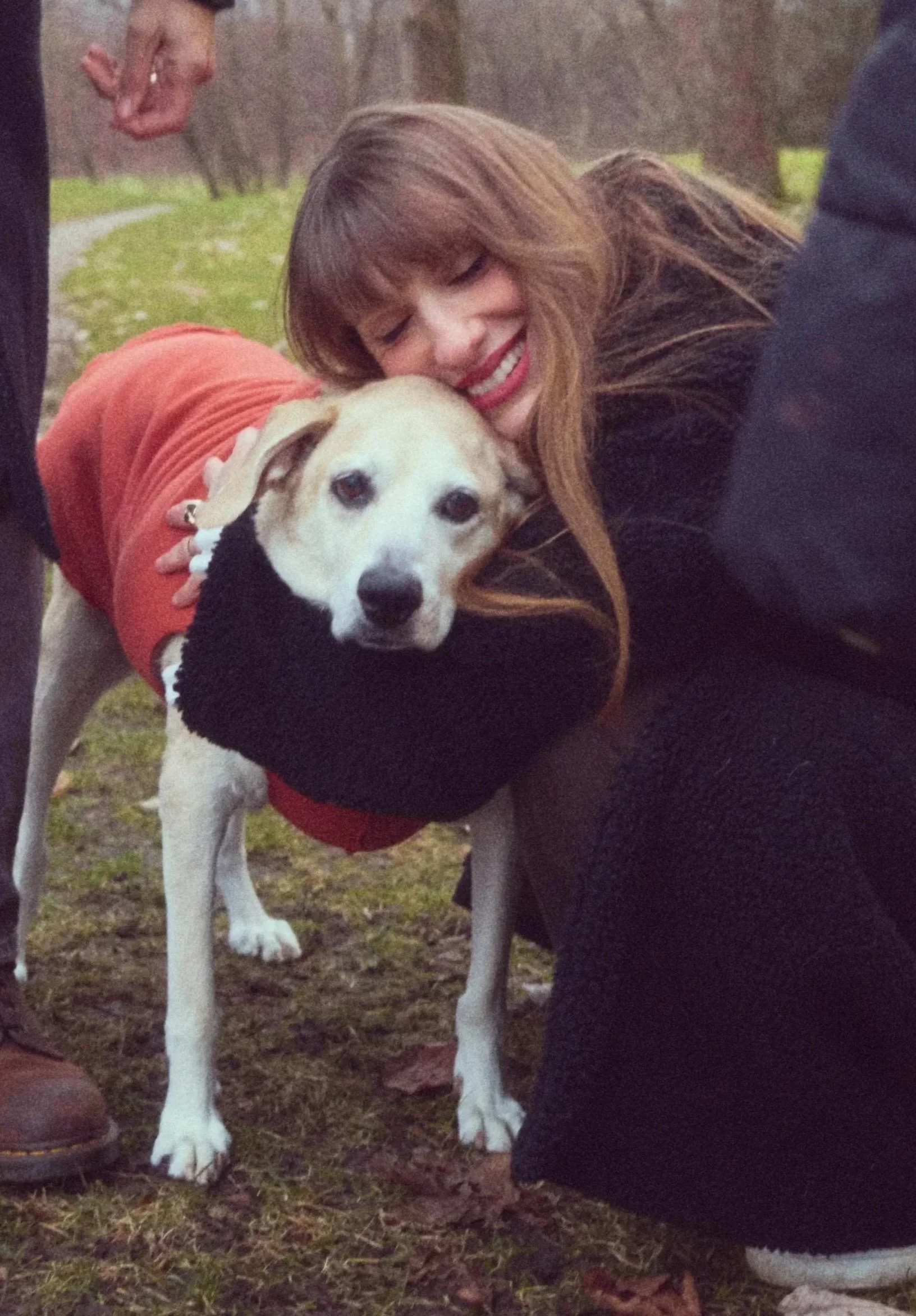 A woman with long brown hair hugging an old dog with tan and white fur, wearing a red jacket. The woman is smiling, and they are outdoors on a grassy area with trees in the background.