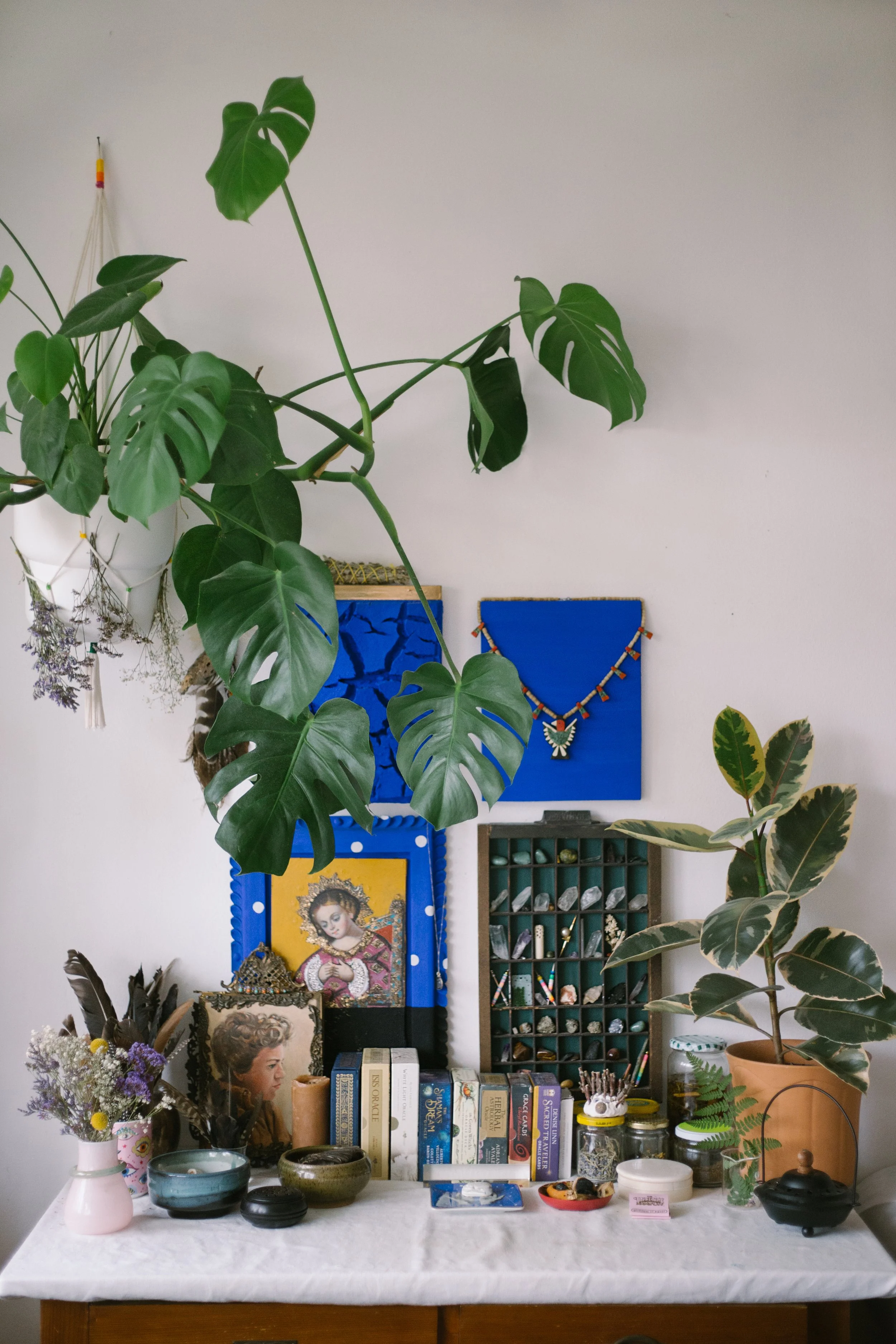 A table with various religious and decorative items, including framed images of Mary and a young woman, books, small bowls, jars, and a potted plant. In the background, there are two framed artworks and a necklace hanging on the wall.