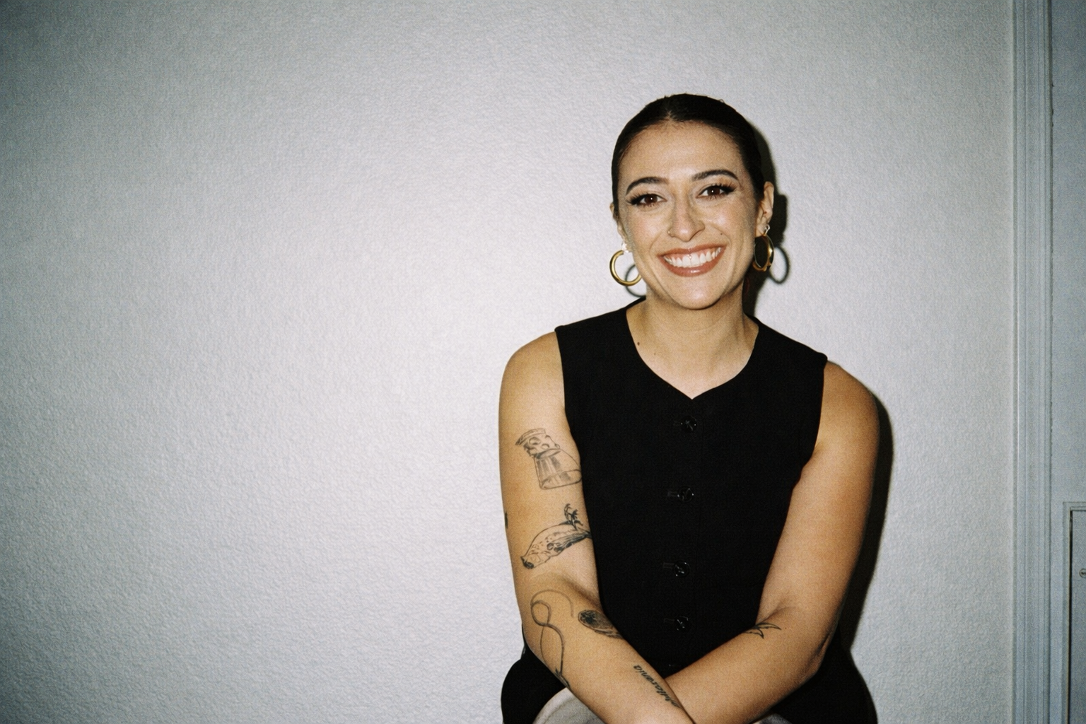 A smiling woman with tattoos on her arms, wearing a black sleeveless top, gold hoop earrings, and makeup, standing against a plain wall.