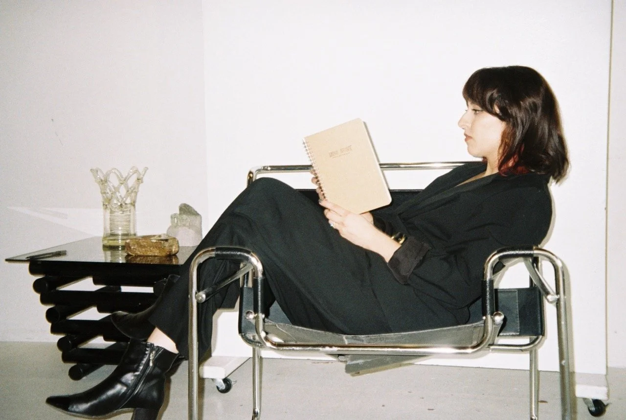 A woman dressed in black, with dark hair, sitting in a modern metal chair, reading a notebook, with a table next to her holding a glass container, a stone, and other items against a plain white wall.