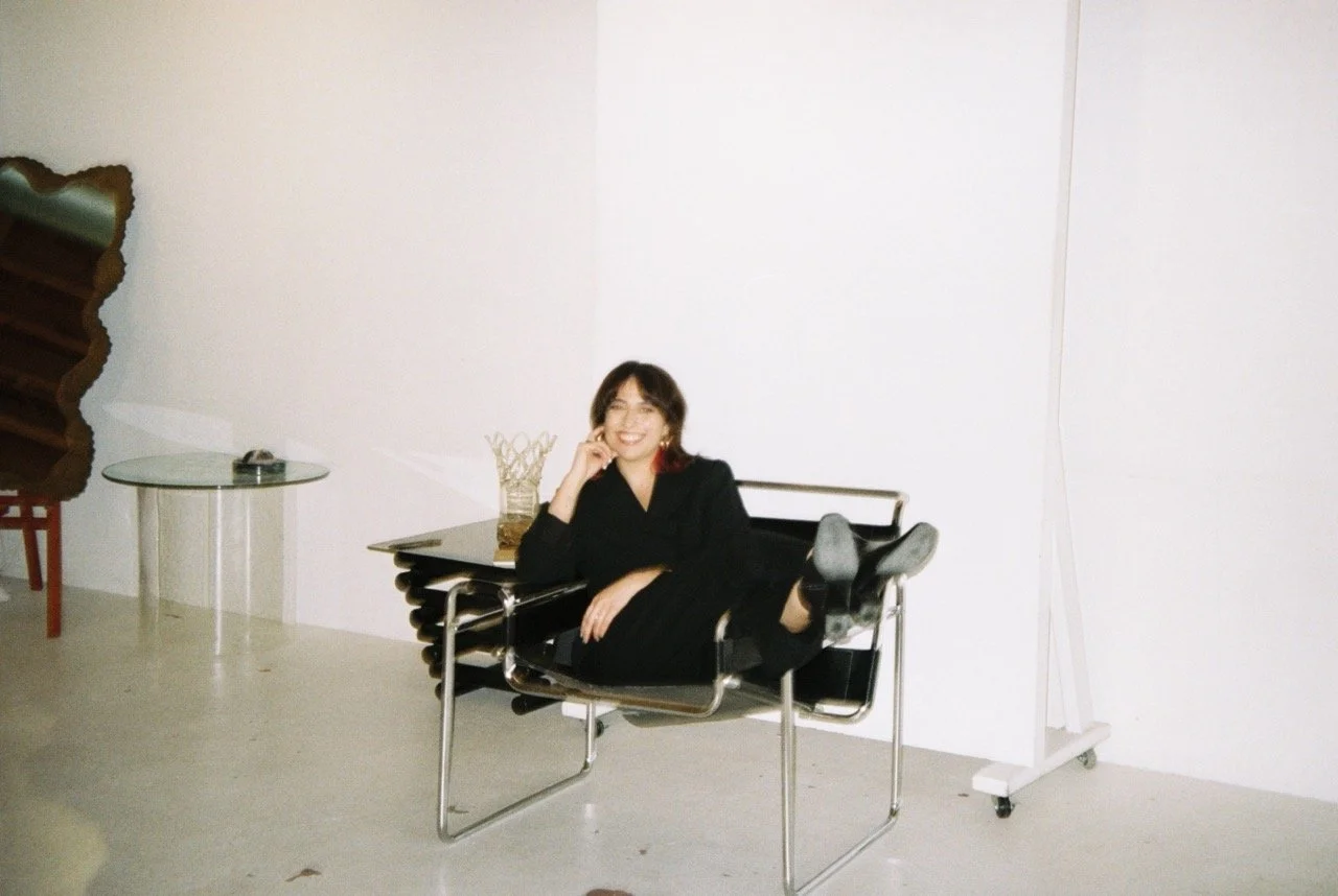 A woman in black clothing reclining and smiling on a modern chair in a minimalistic room with white walls, a glass table, and a large whiteboard on wheels.