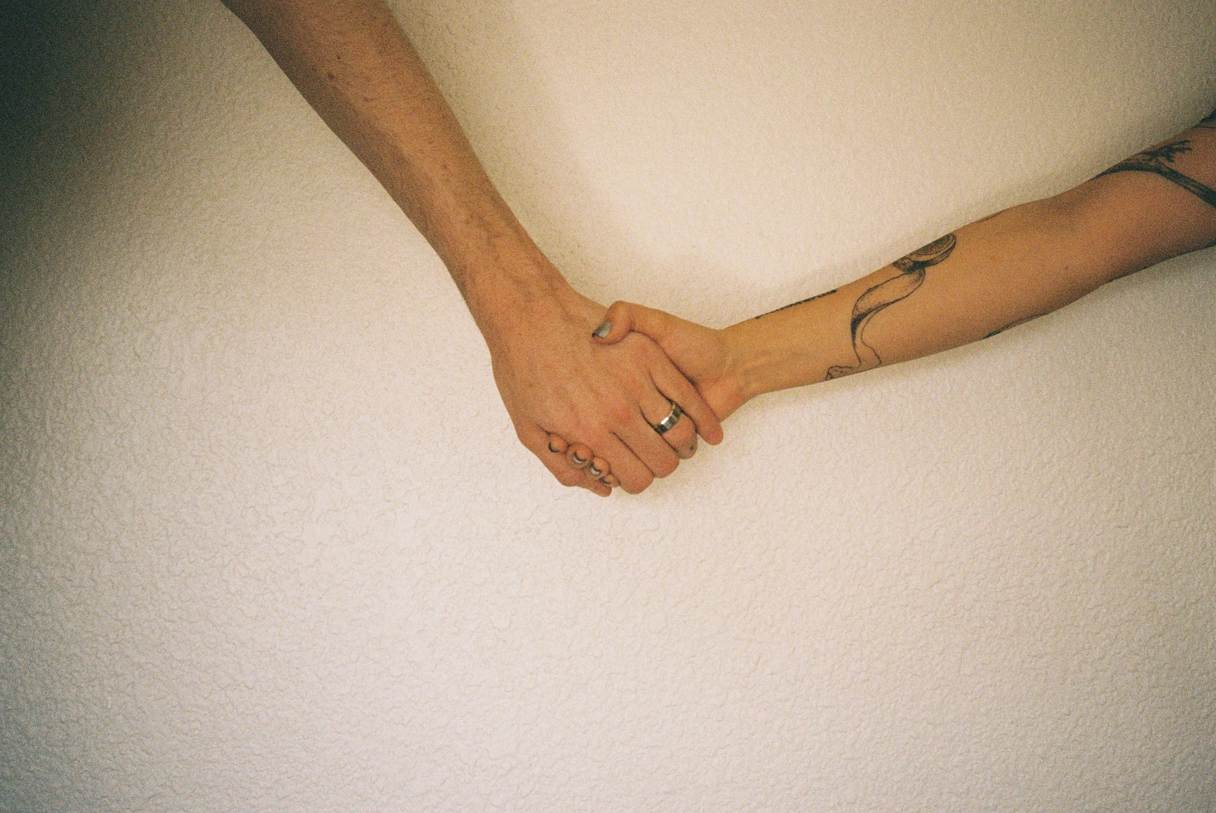 Two people holding hands against a plain wall, with one person's arm featuring a tattoo of a snake.