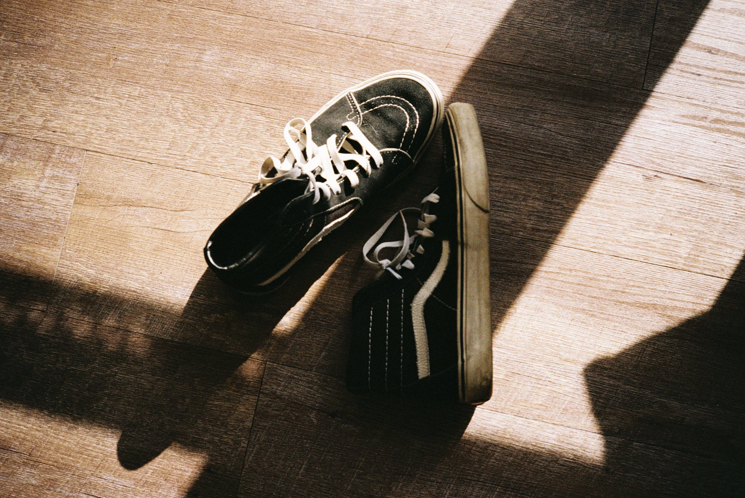 Pair of black and white sneakers on wooden floor, with sunlight casting shadows.